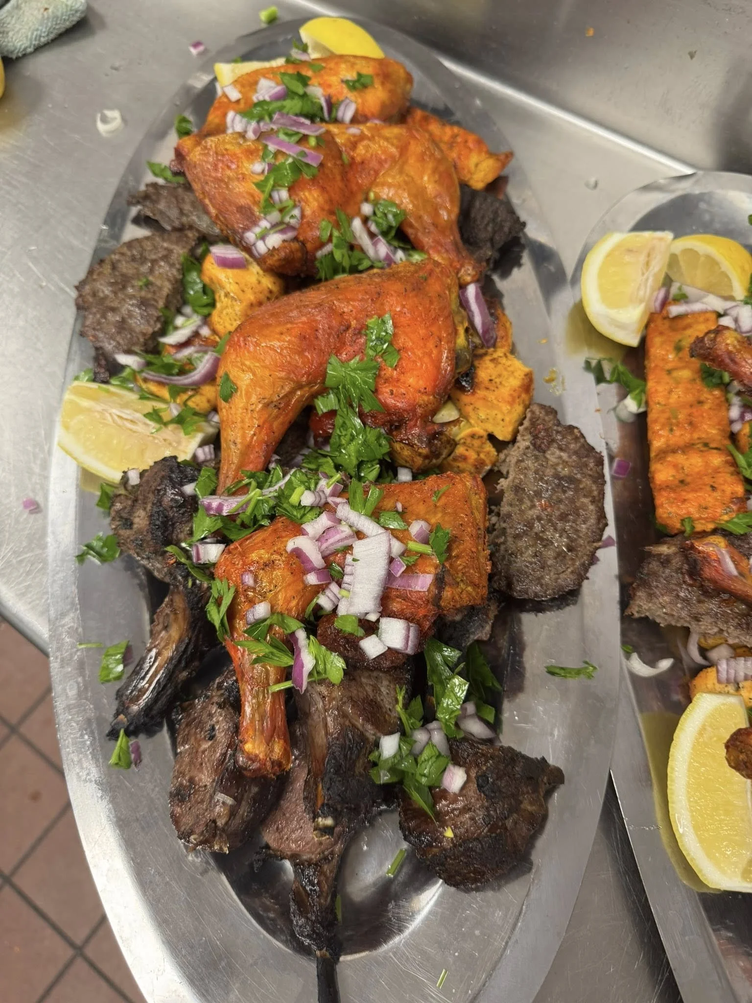 A platter of grilled meats including lamb, beef, and chicken, garnished with chopped red onions, cilantro, and lemon wedges.