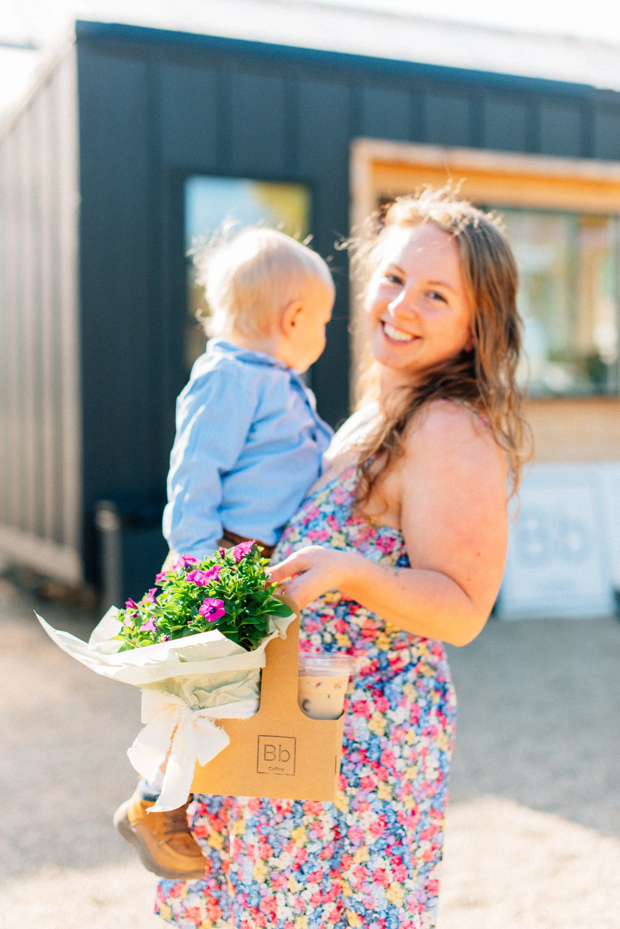 A woman with long, curly hair smiling while holding a young child in one arm and a bouquet of pink flowers wrapped in brown paper in the other hand.