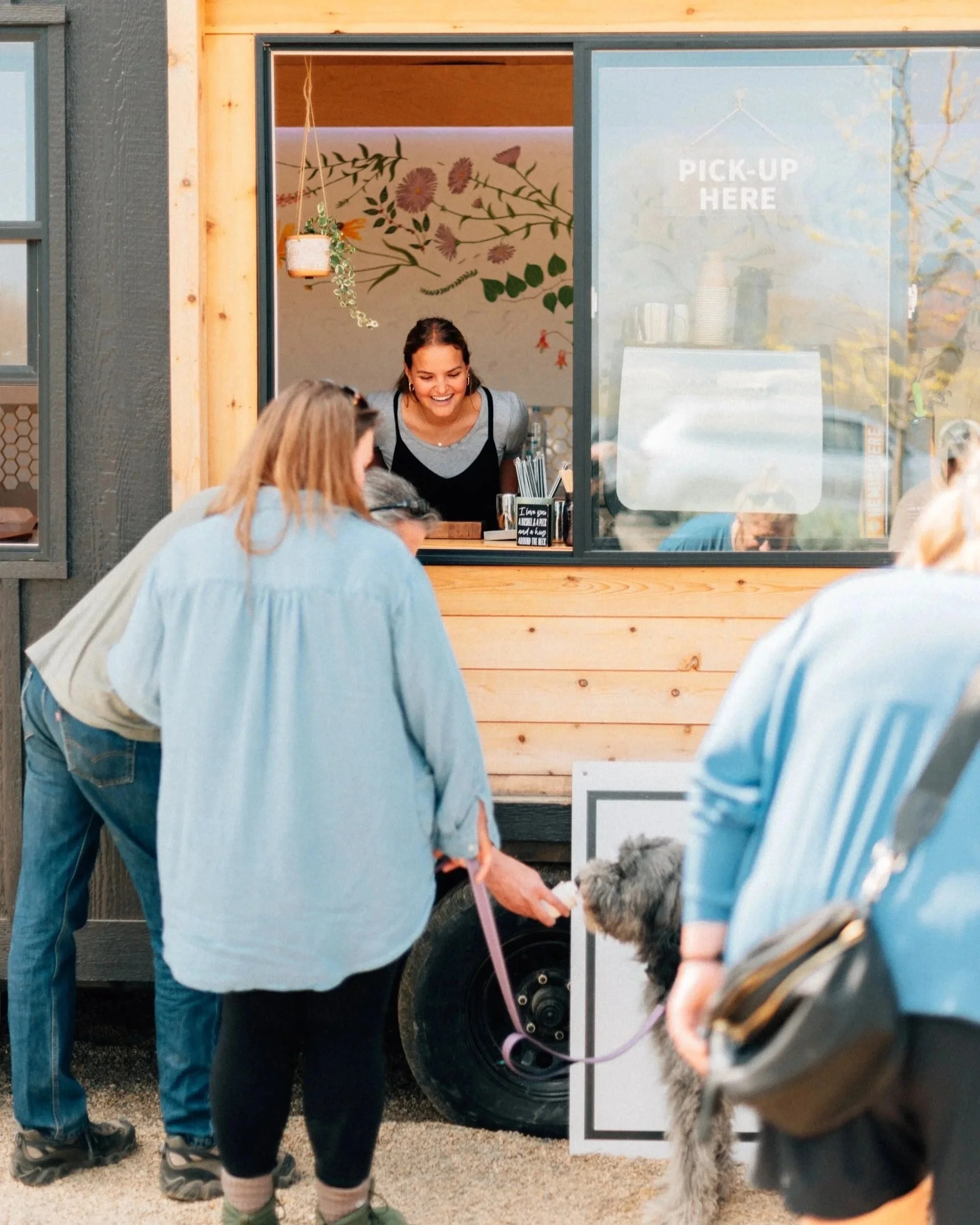 People ordering food at a food truck window with a woman inside smiling, and a dog being pet by a person in front of the truck.