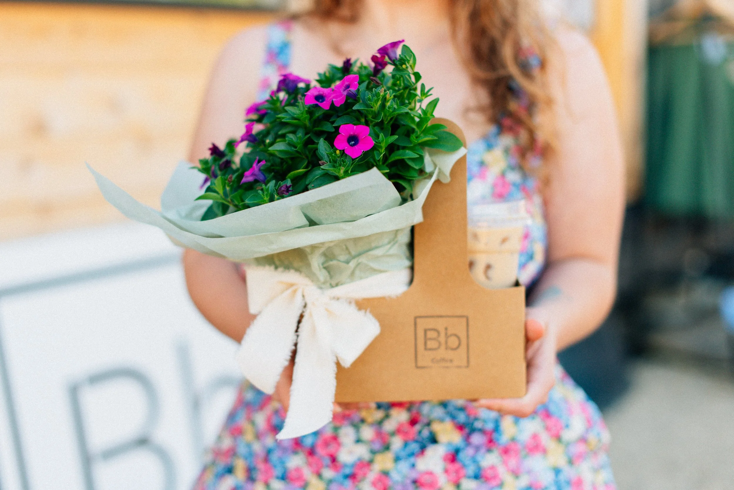 A young girl holding a potted plant with pink and purple flowers wrapped in white paper and placed inside a cardboard box.