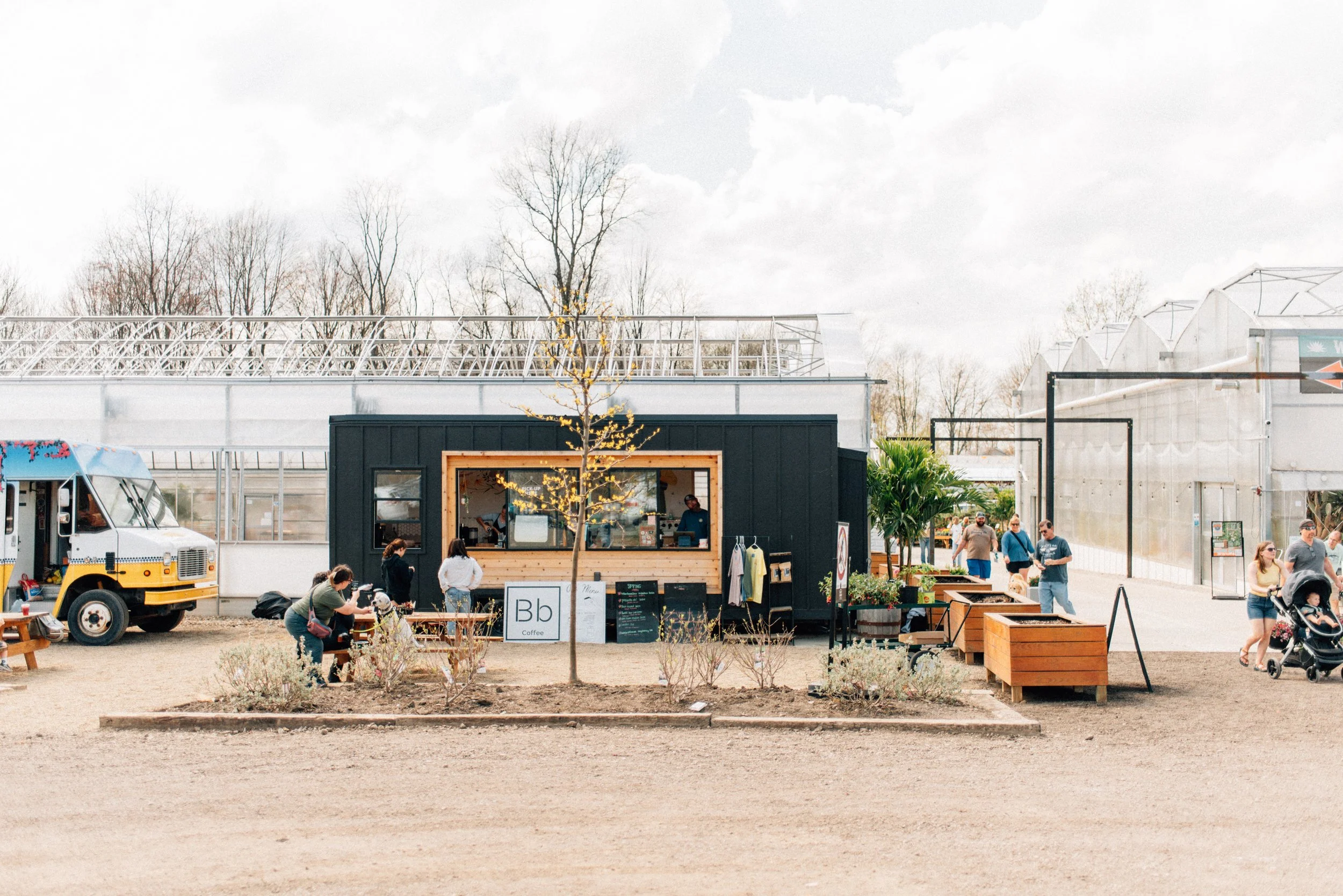 A small black coffee stand with a menu board outside, surrounded by a few people, and a yellow food truck on the left. There are trees and greenhouses in the background, with some clouds in the sky.