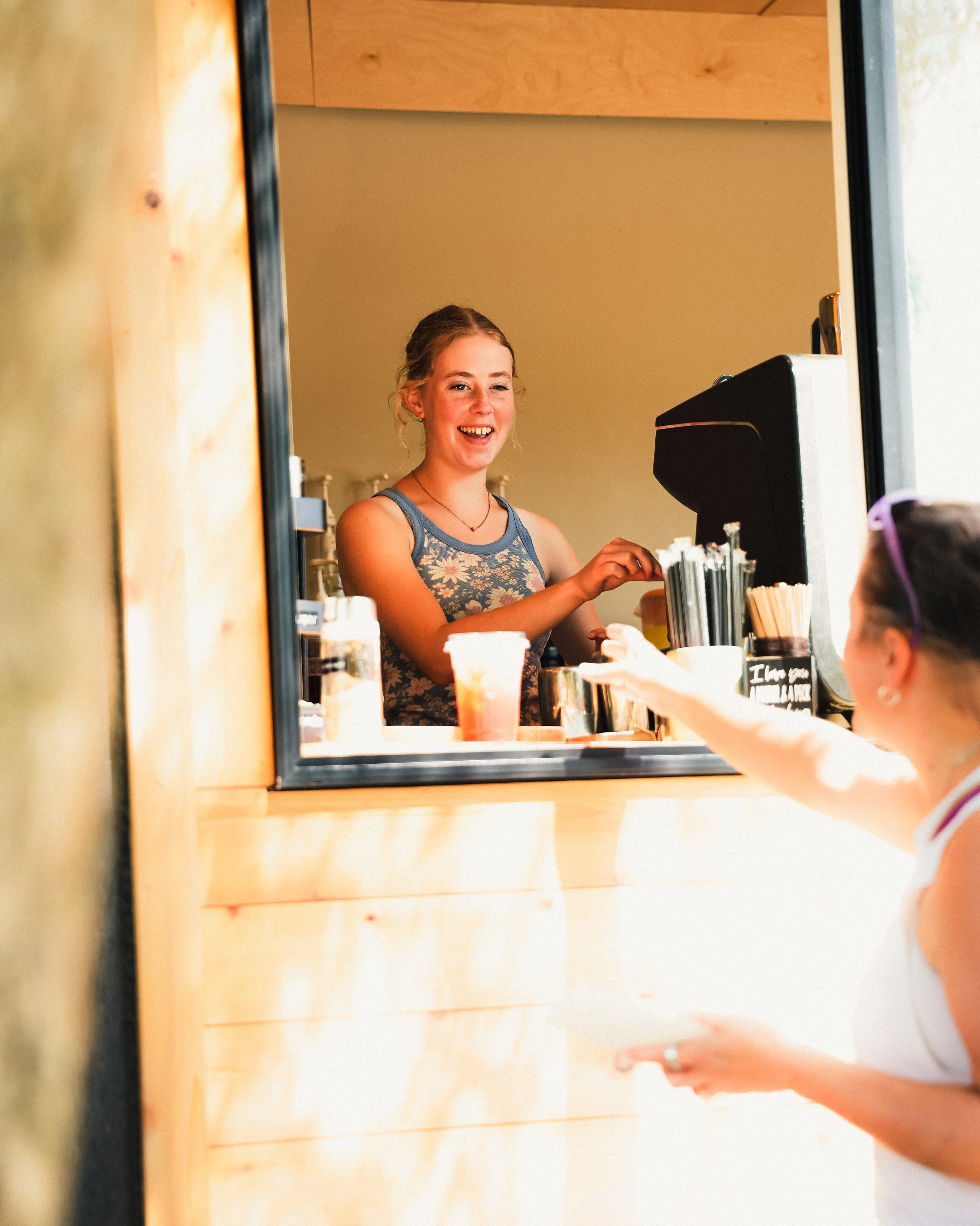A young girl with braided hair and a floral dress is smiling while ordering at a coffee stand through a window, reaching out with money or a card to pay.