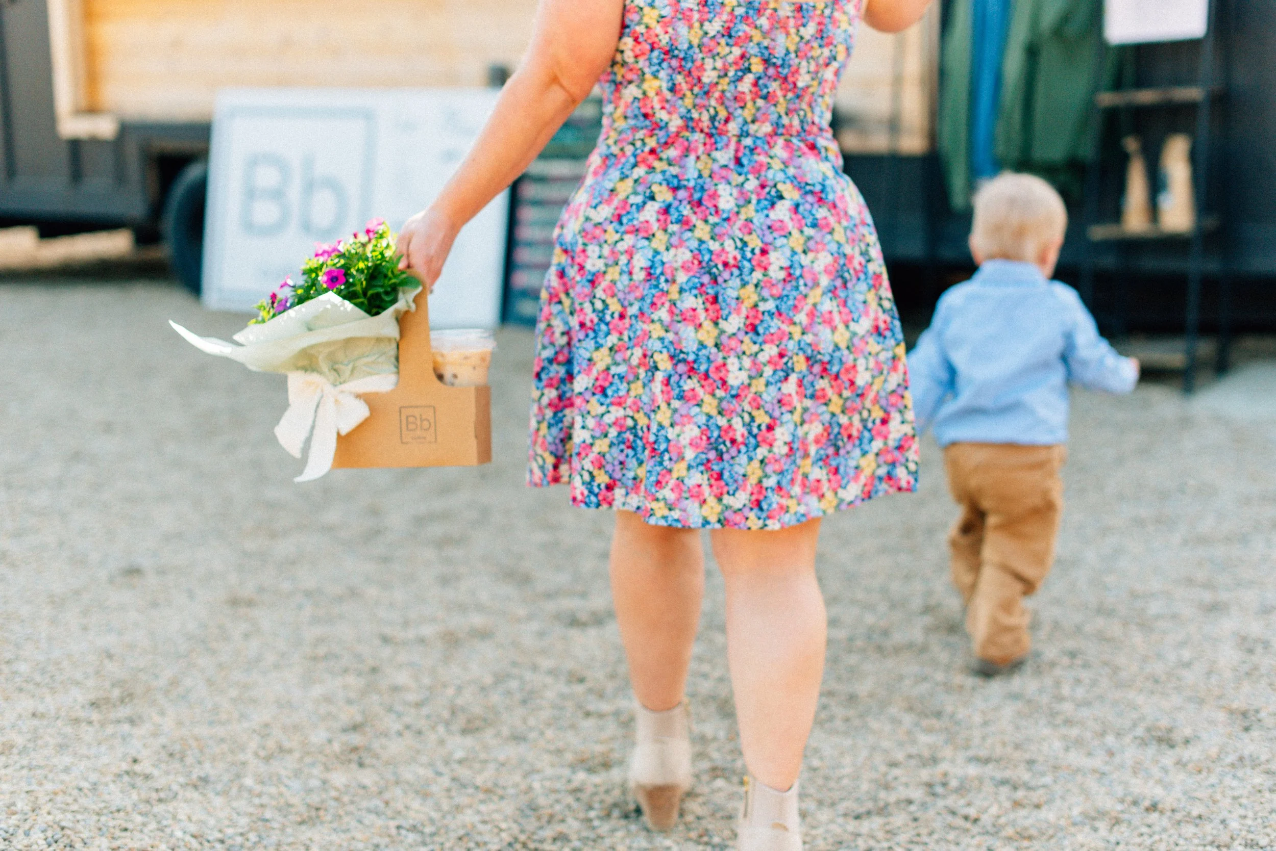 A woman in a colorful floral dress holding a paper bag with flowers and a drink, walking on a gravel path with a child in a blue shirt and brown pants walking ahead.