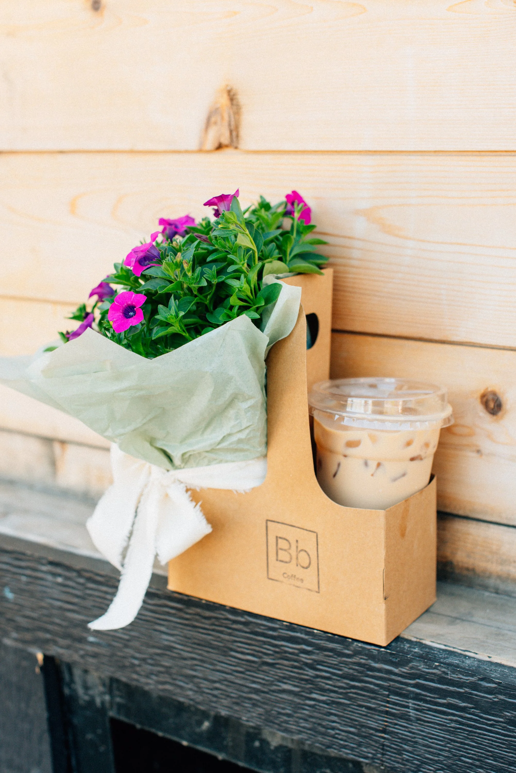 A cardboard box containing a potted plant with purple and pink flowers and a small plastic cup of iced coffee on a dark wooden outdoor table.