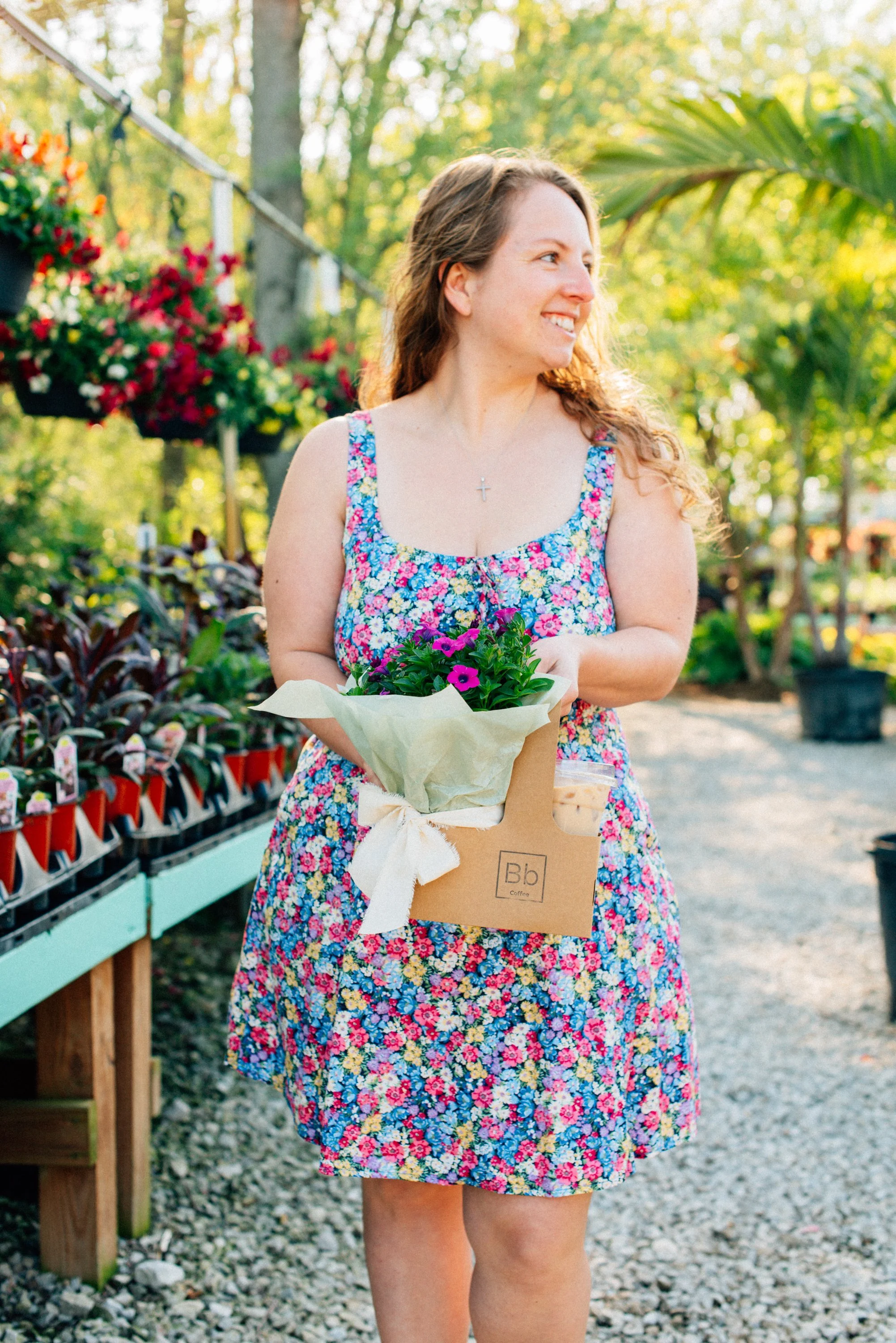 A woman holding a small bouquet of pink and purple flowers, wearing a floral dress and a cross necklace, standing outdoors at a garden center or nursery with hanging flower baskets and potted plants in the background.