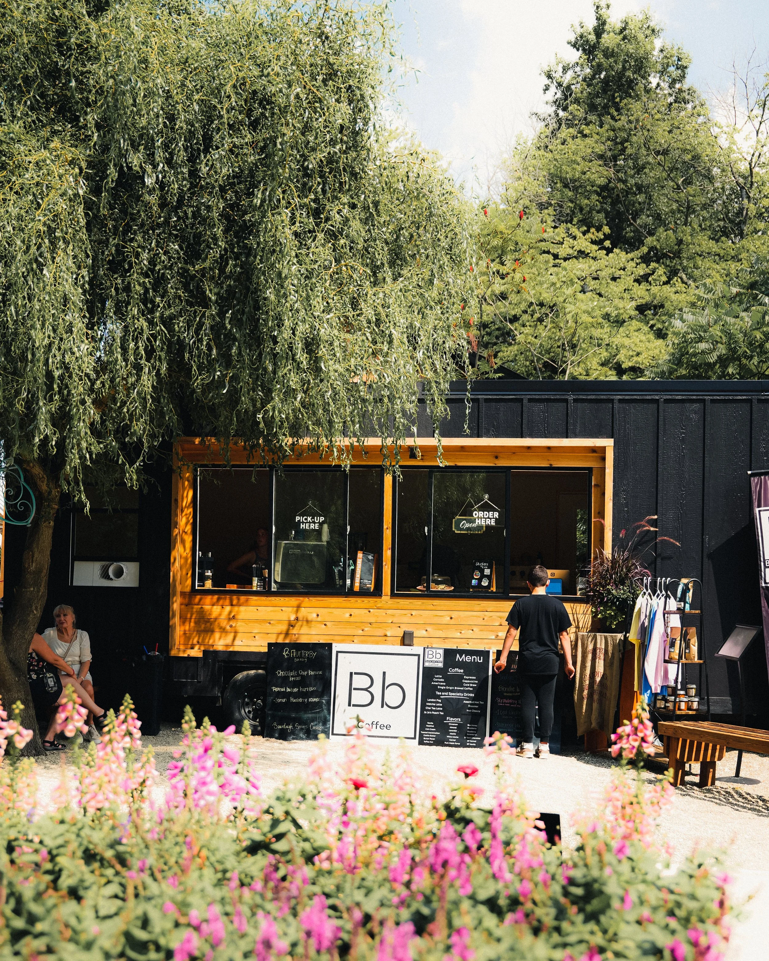 A small coffee stand with a black exterior and wooden accents, serving customers outdoors on a sunny day, with a flower bed in the foreground and trees in the background.