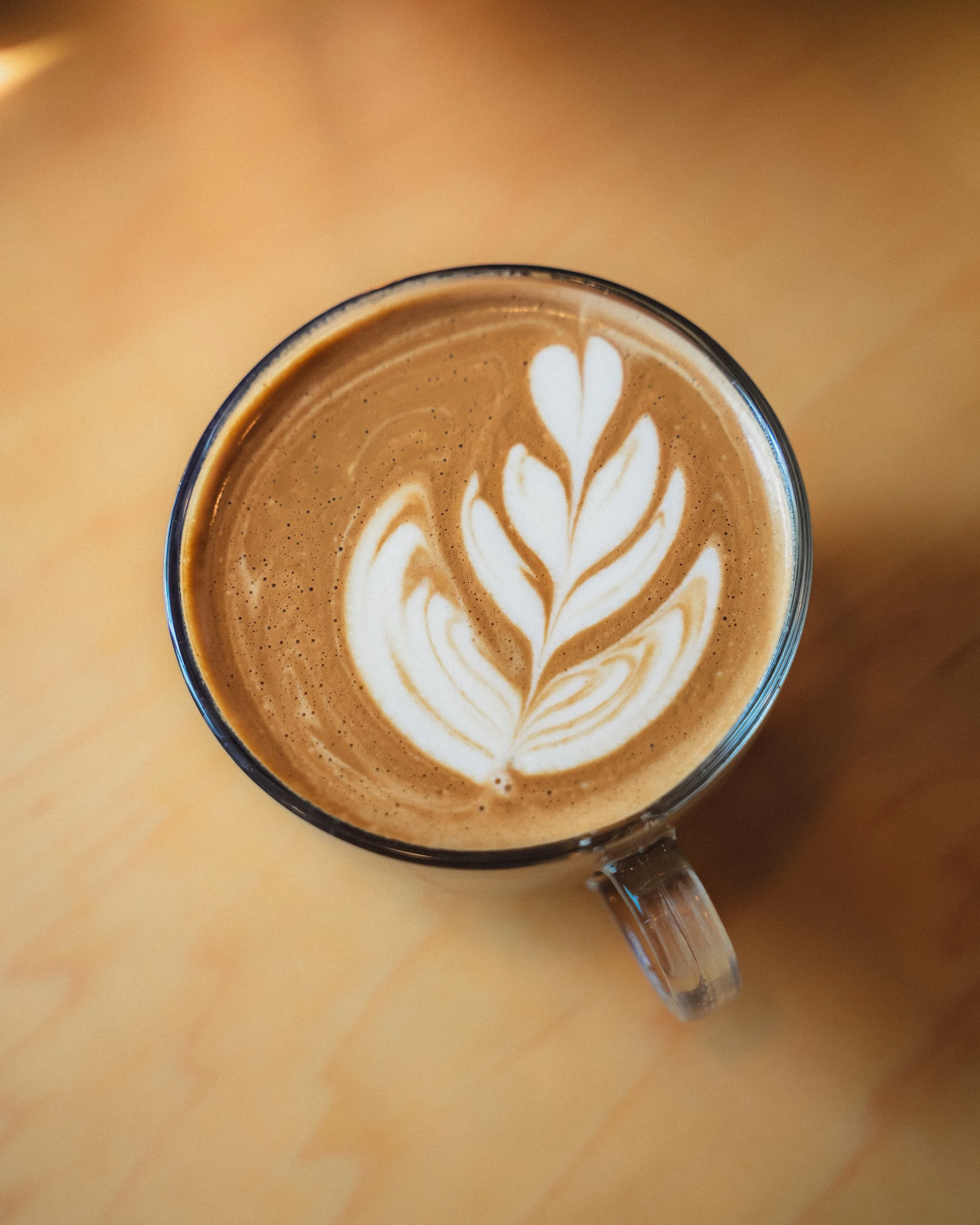 A top-down view of a glass cup filled with a latte topped with white foam art resembling a floral pattern.
