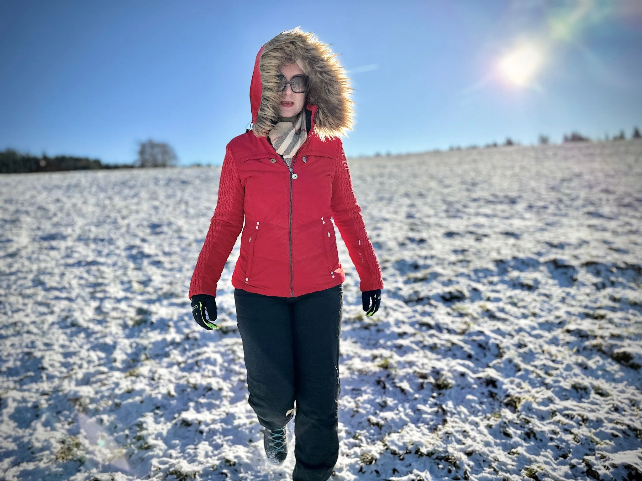 Auf einer Winterwanderung im Val de Travers mit Winterjacke und Kapuze läuft Joëlle Gautier auf verschneitem Feld bei Sonnenschein.