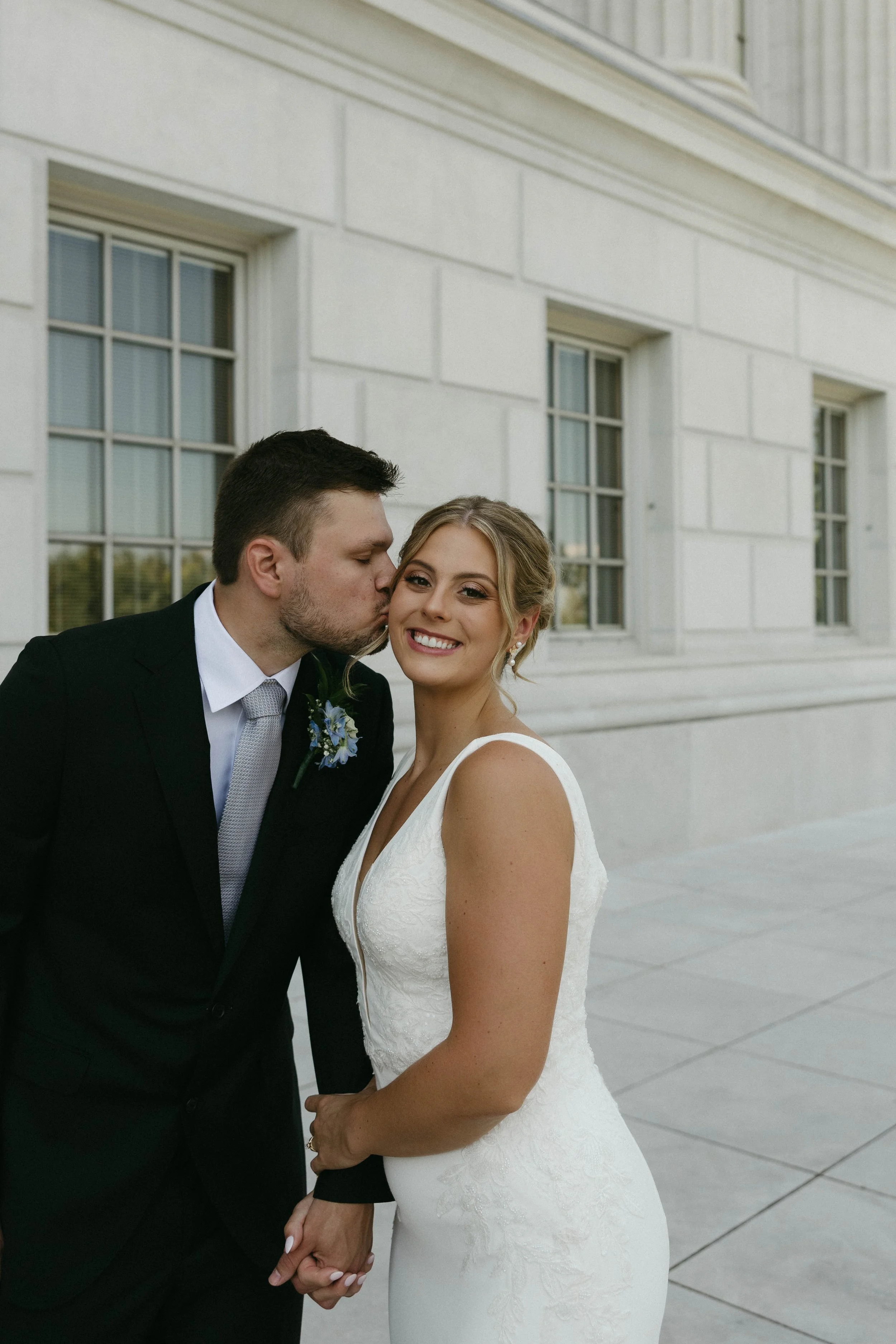 A newlywed couple holds hands, with the groom giving the bride a gentle kiss on the cheek. The bride is smiling, wearing a white wedding gown, and the groom is dressed in a black suit with a light gray tie. They are standing outside in front of a cream-colored building with large glass window panes.