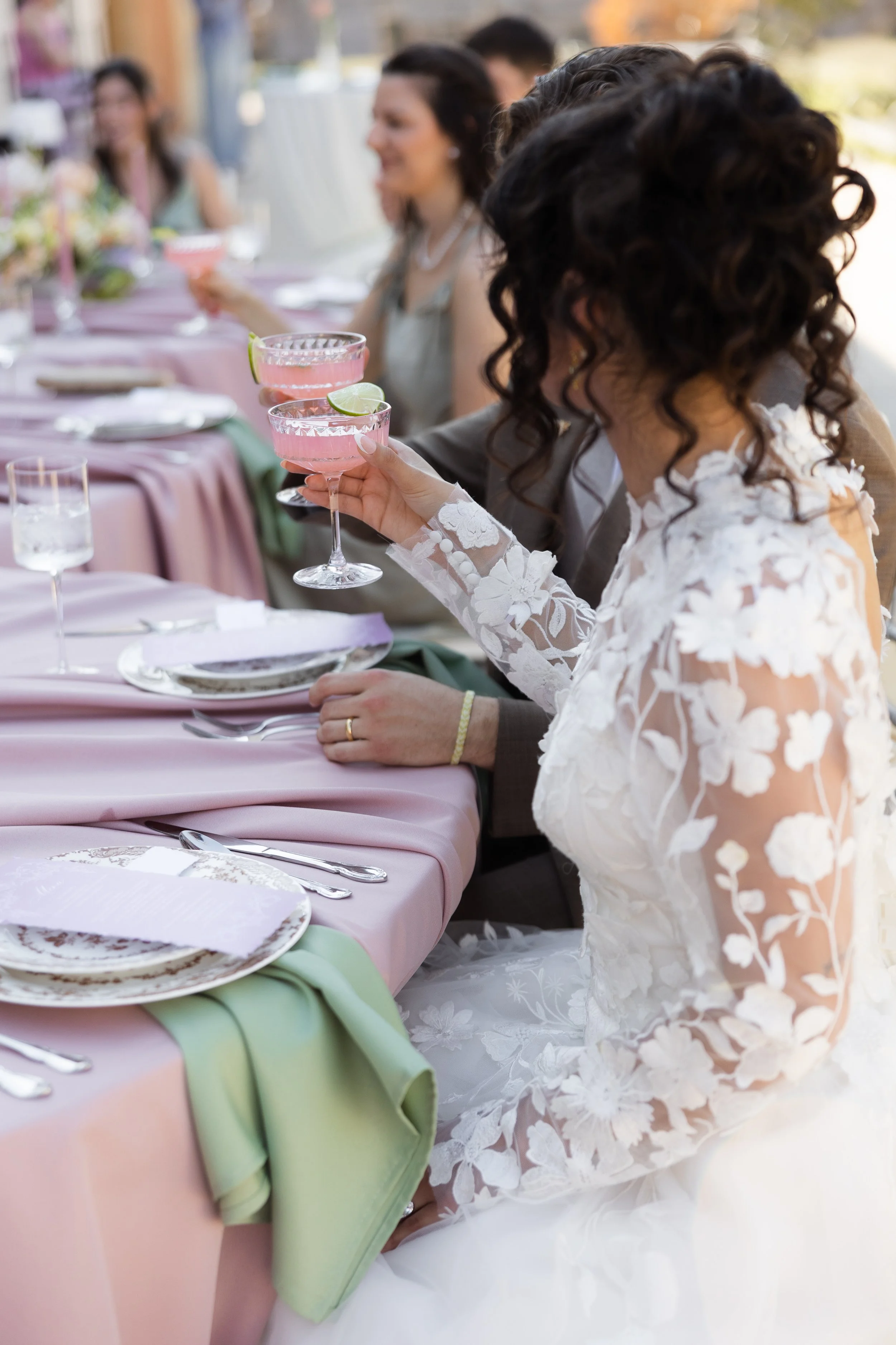 Close-up of a woman with curly hair wearing a white lace wedding dress, holding a pink cocktail garnished with a lime wedge, at a formal celebration table decorated with pastel tablecloth, plates, and cutlery.