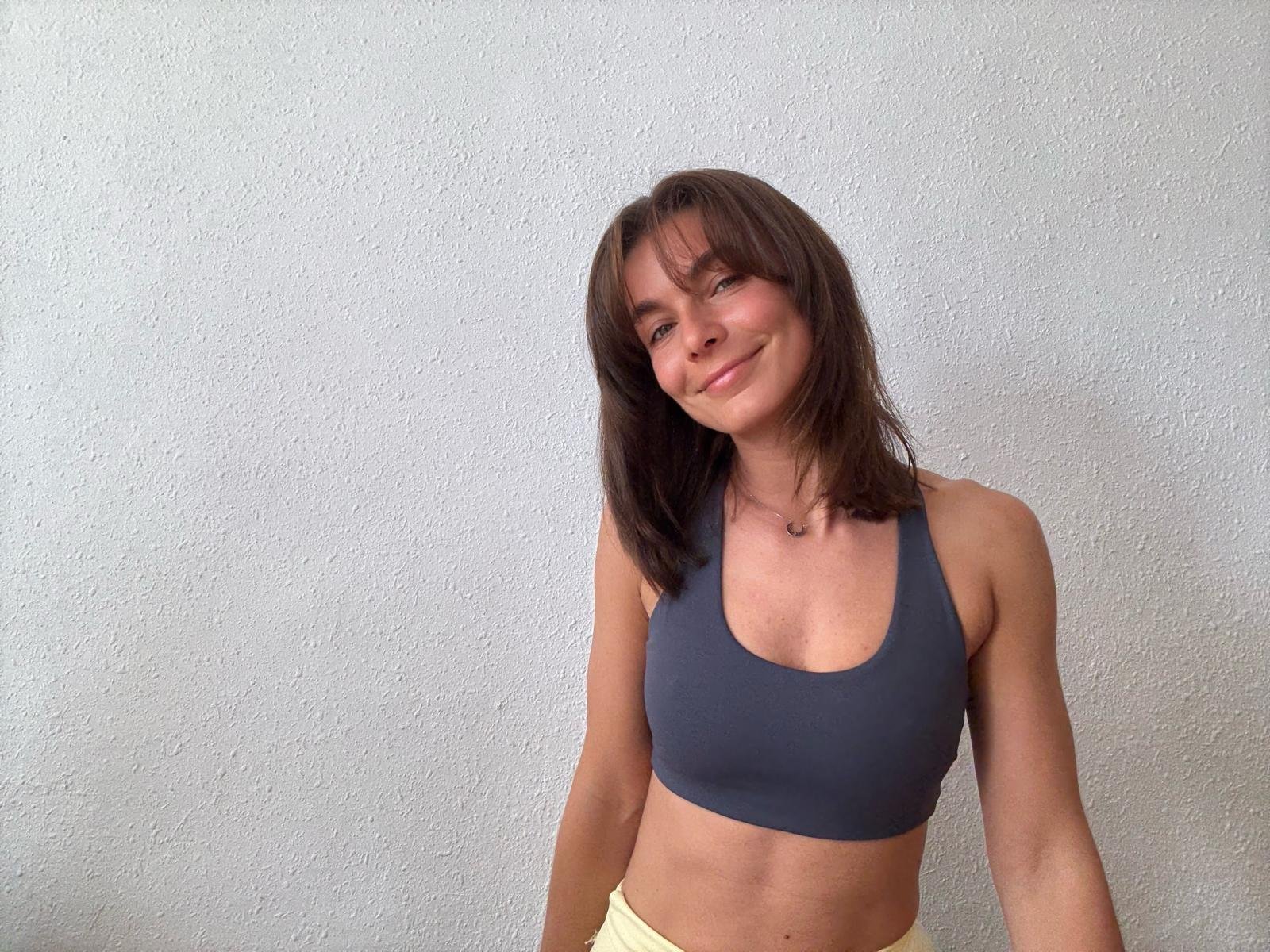 A young woman with shoulder-length brown hair, smiling and winking, dressed in a dark blue sports bra and yellow shorts, standing against a white textured wall.