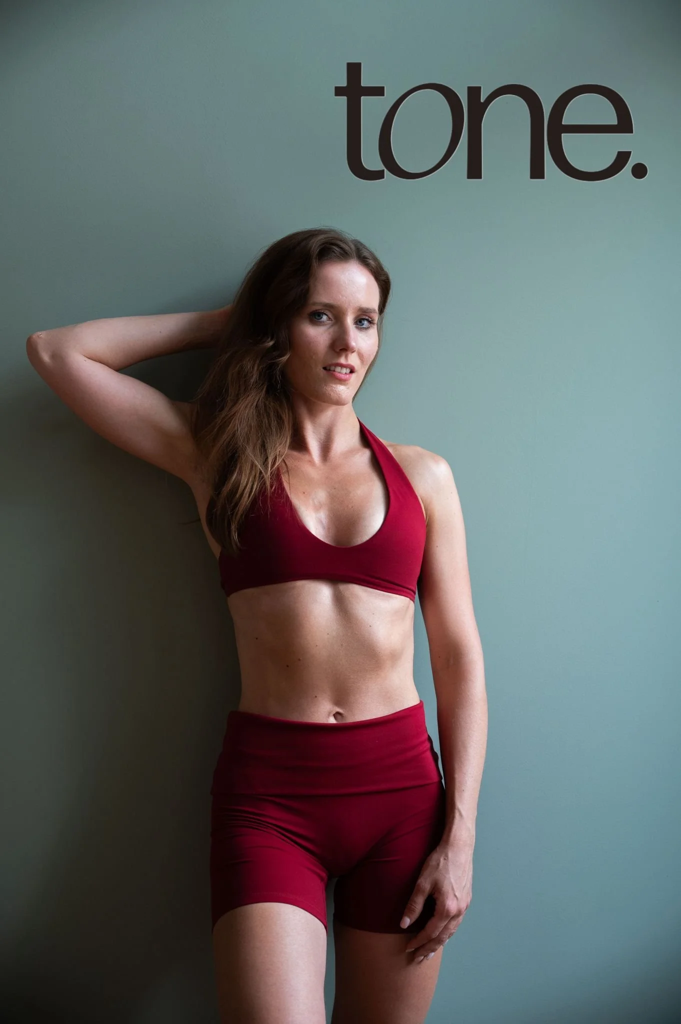 Žena v červeném sportovním oblečení pózuje u zelené zdi s černým nápisem „tone.“.