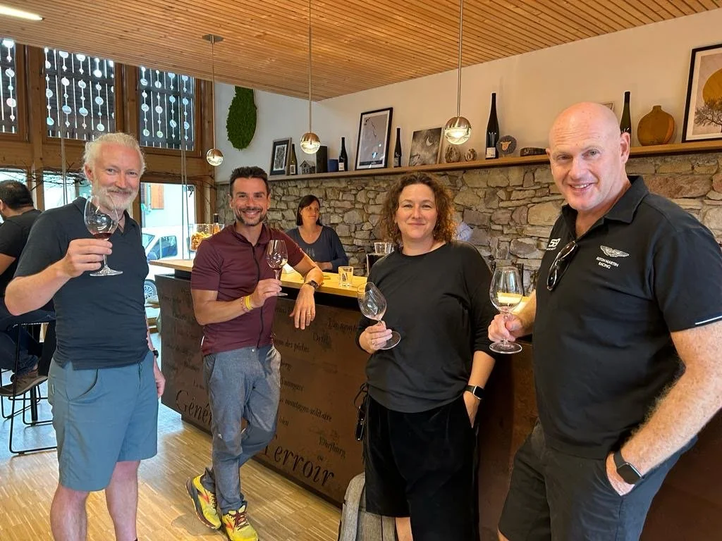 Four people standing inside a rustic restaurant, holding wine glasses and smiling at the camera. The background features a stone wall, wooden accents, and decorative bottles and framed pictures on a shelf.