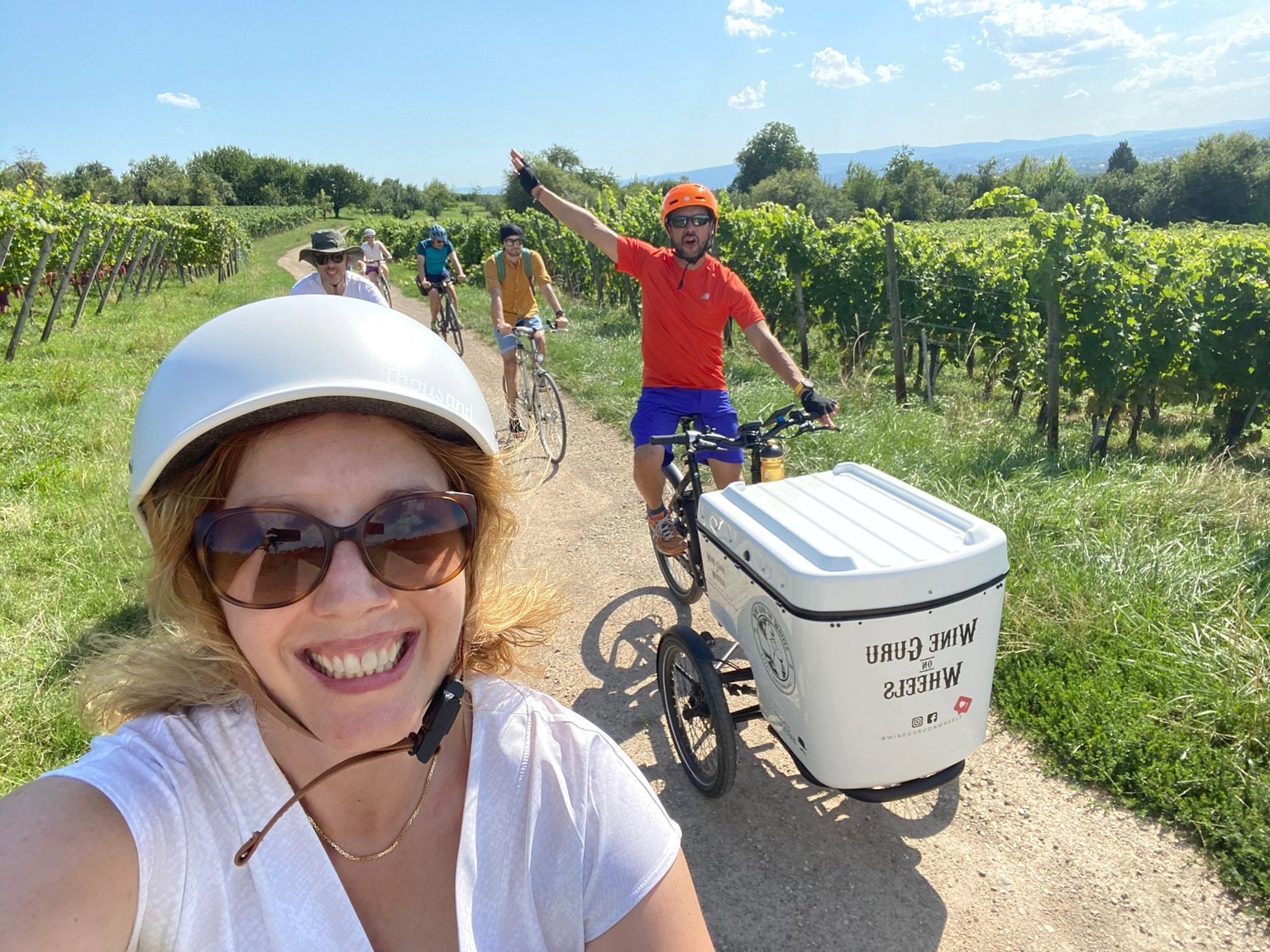 A group of people riding bicycles on a dirt path through a vineyard, with a woman in the foreground smiling and wearing sunglasses and a helmet, and a man in a red shirt and helmet pointing ahead with a bike trailer in front of him.