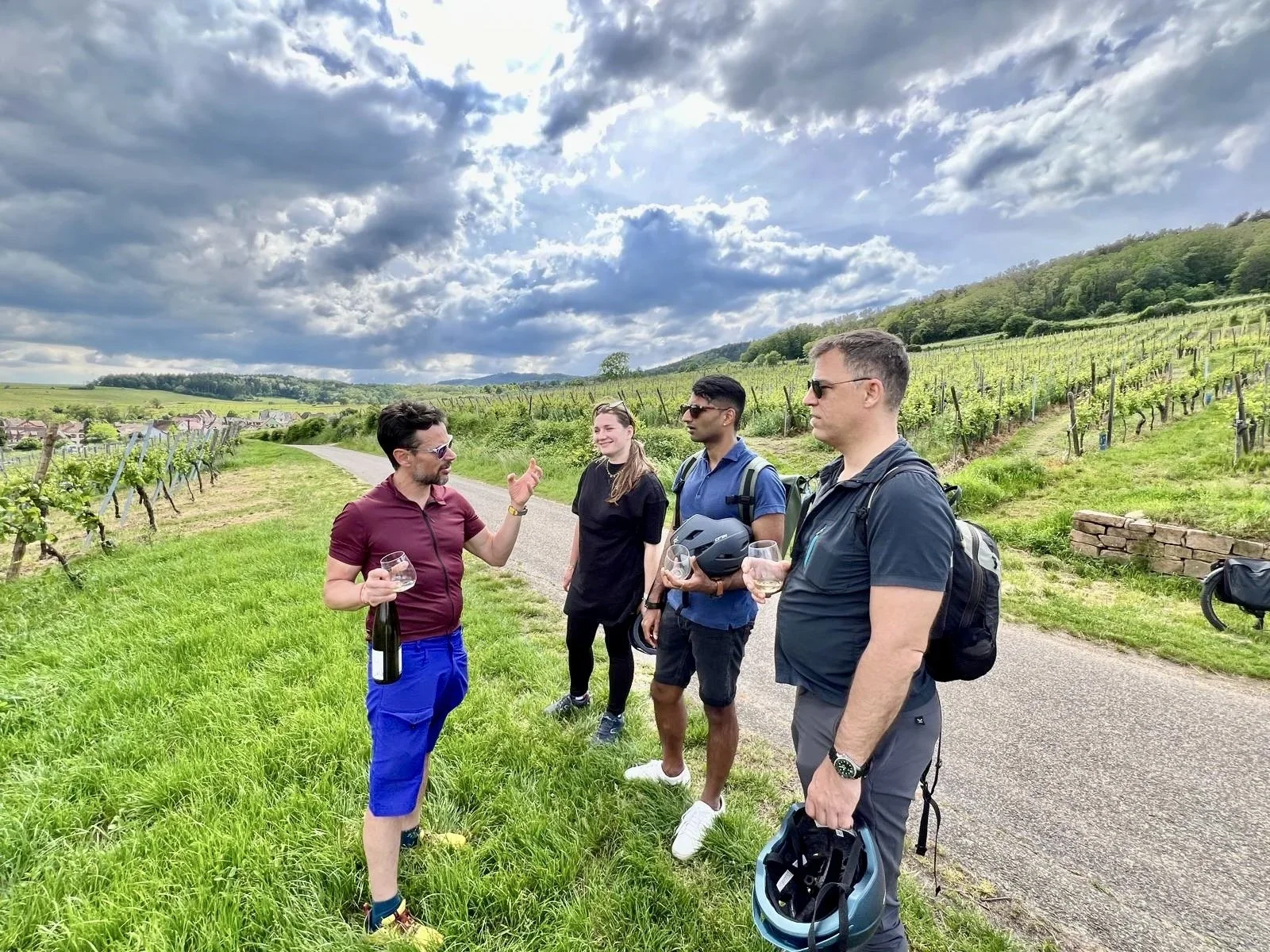 Group of four people having a conversation in a vineyard, two men in sunglasses holding glasses of wine, one man holding a helmet, the woman smiling, behind them green vineyards and a cloudy sky.