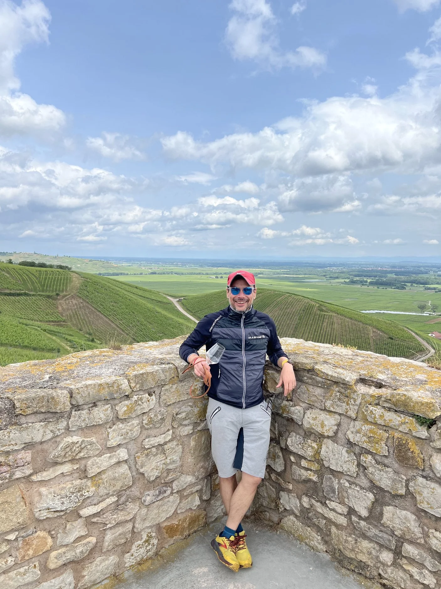 A man standing on a stone observation point overlooking lush green rolling hills and vineyards under a partly cloudy sky.