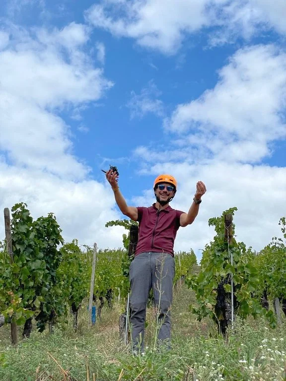 A man in a maroon shirt, gray pants, and orange helmet standing in a vineyard, holding a drone in one hand and raising his other arm, smiling with clouds in the sky behind him.