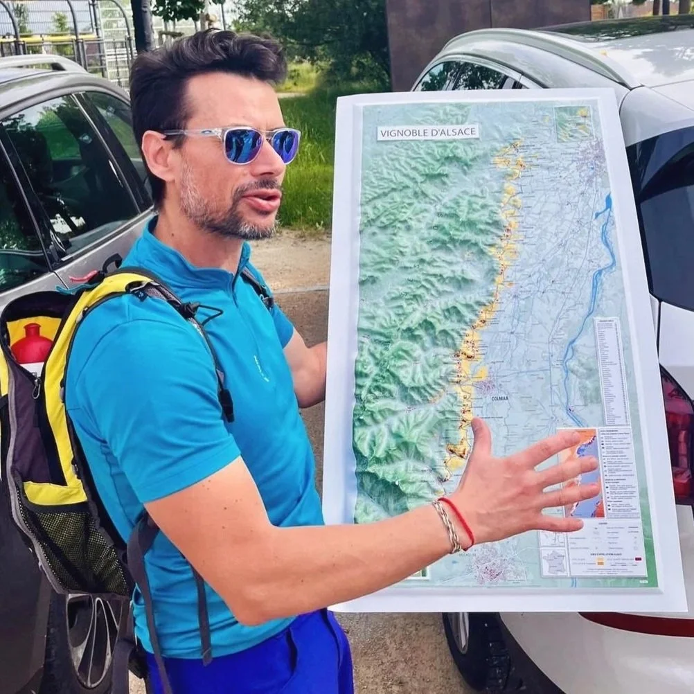 A man in a blue shirt and sunglasses holding a large map of the Alsace region in France near parked cars outdoors.