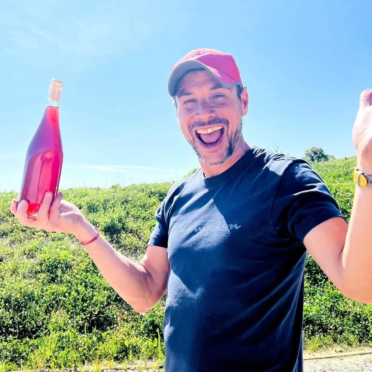 A man wearing a red cap and black t-shirt smiling and holding a bottle of pink-colored drink outdoors on a sunny day, with green bushes and blue sky in the background.