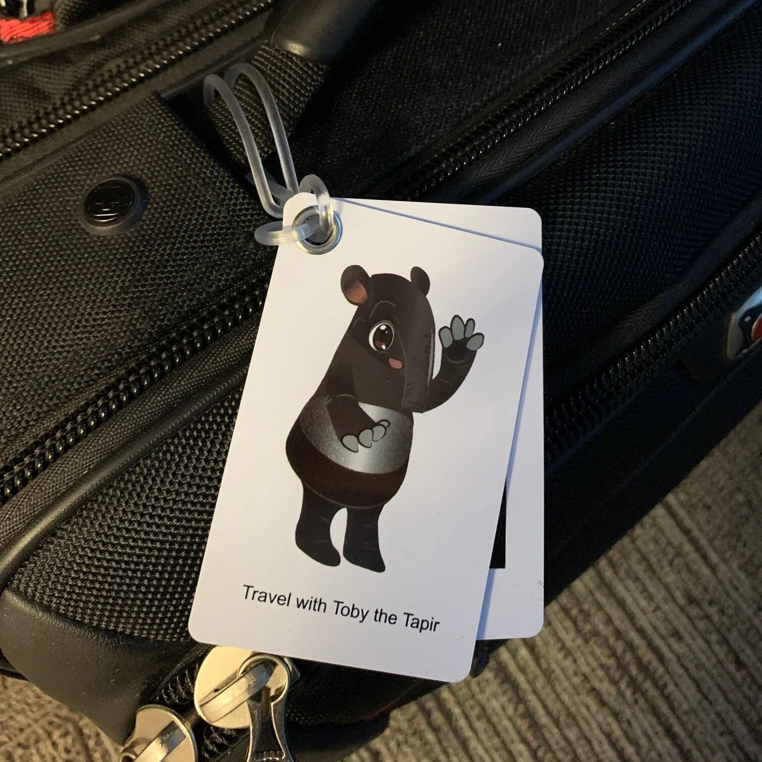 A luggage tag attached to a black suitcase features a cartoon tapir character named Toby, who is waving. The tag includes the text "Travel with Toby the Tapir".