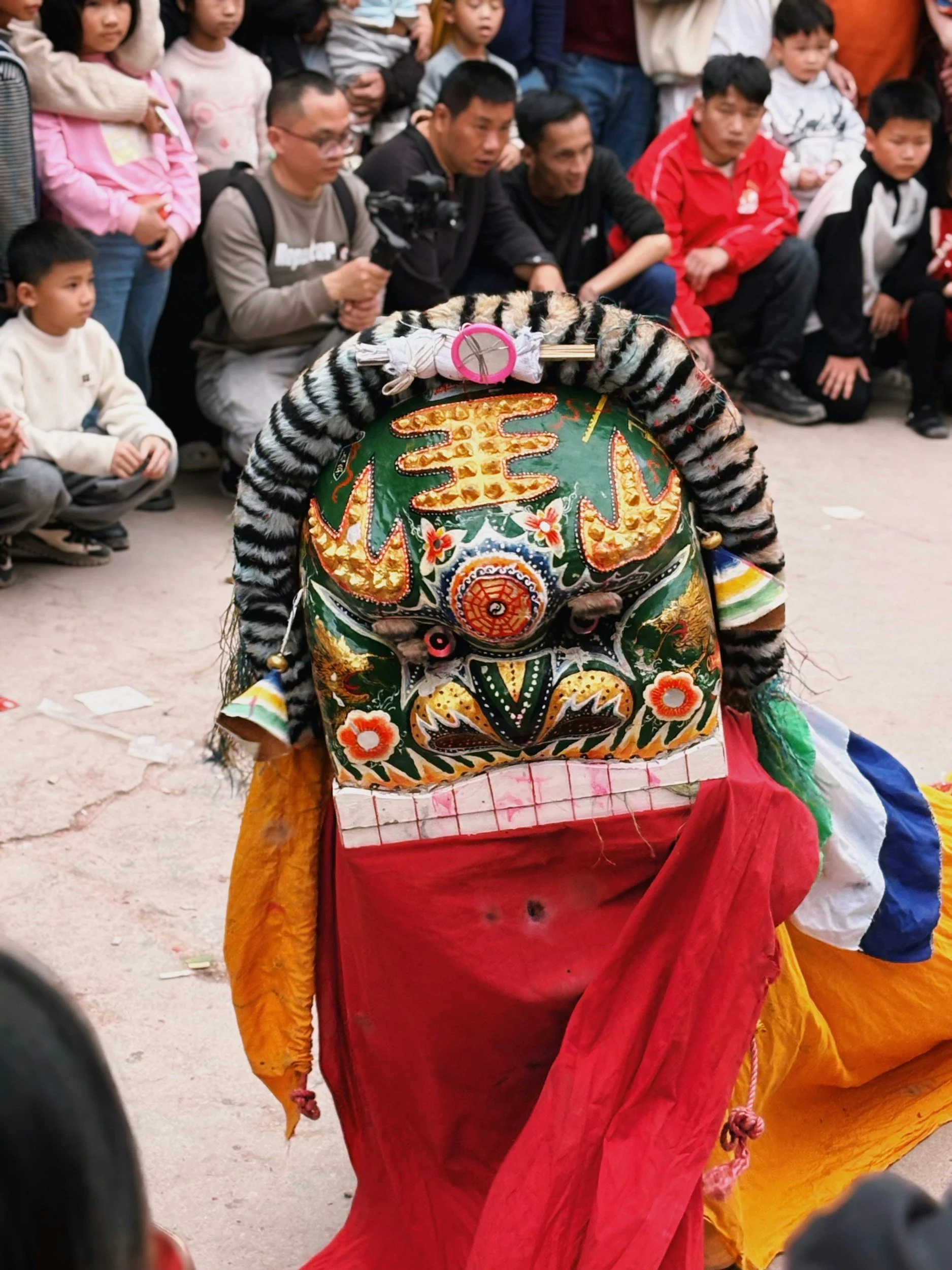 A traditional lion dance costume is seen from behind, featuring colorful, ornate fabric with floral and circular patterns, as performers wear tiger and lion costumes while an audience of children and adults watch in the background.