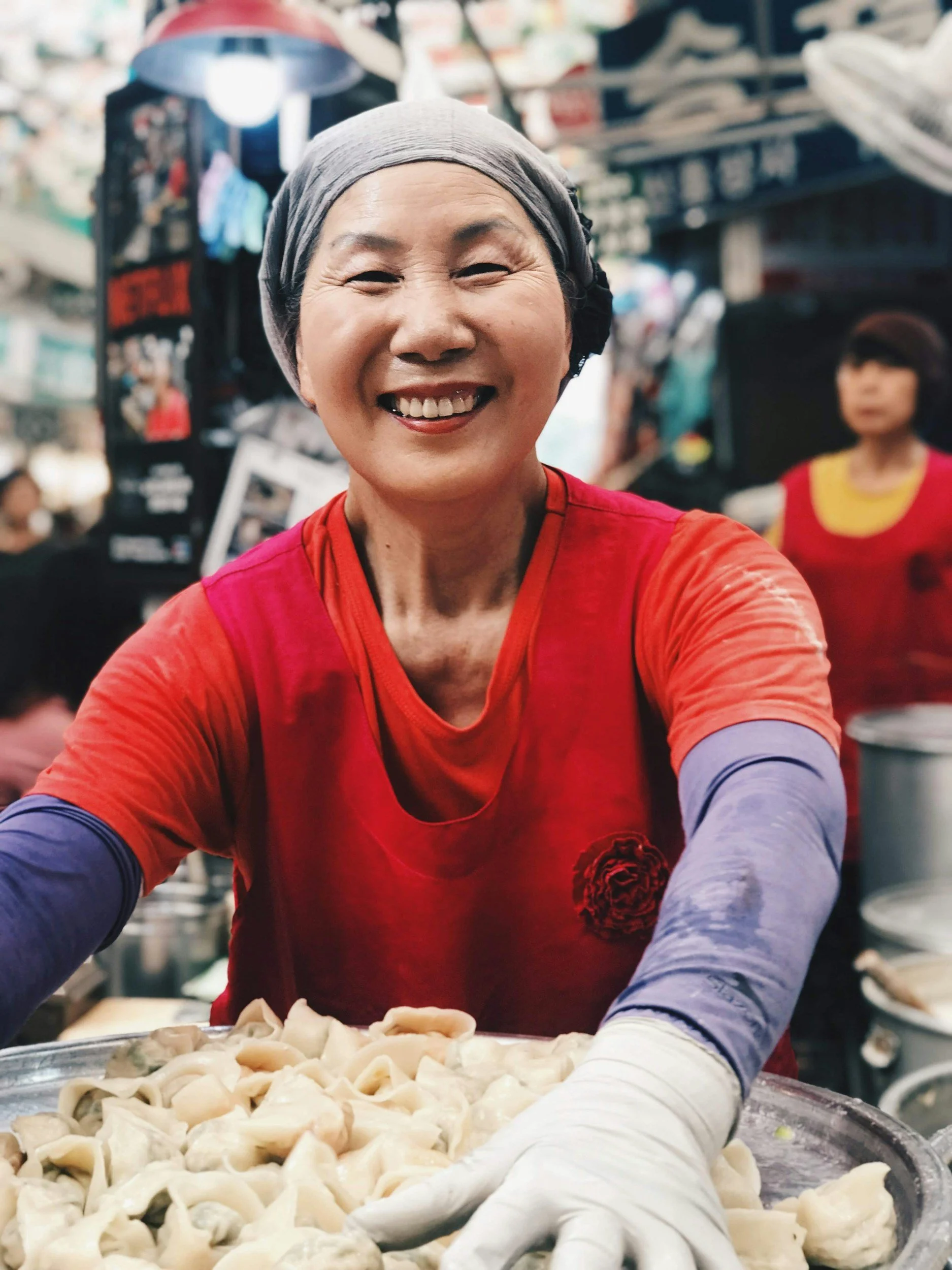 Smiling woman wearing a gray headscarf and red shirt, working in a crowded market. She is making dumplings, with a large tray of dough in front of her.