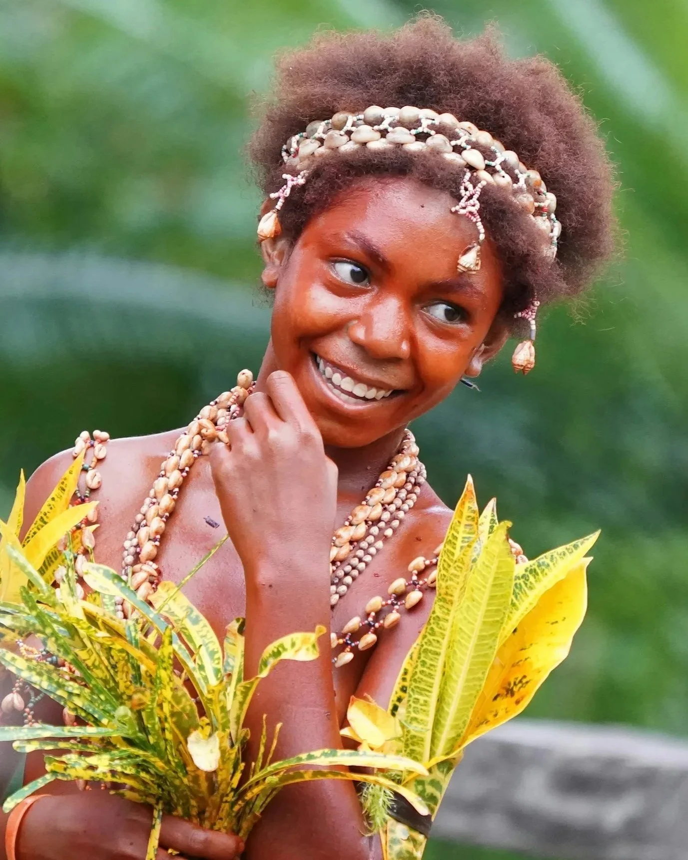 A woman with dark skin and short, curly hair wearing traditional jewelry with shells, and a yellow patterned dress, holding green and yellow tropical plants while smiling outdoors against a green blurred background.