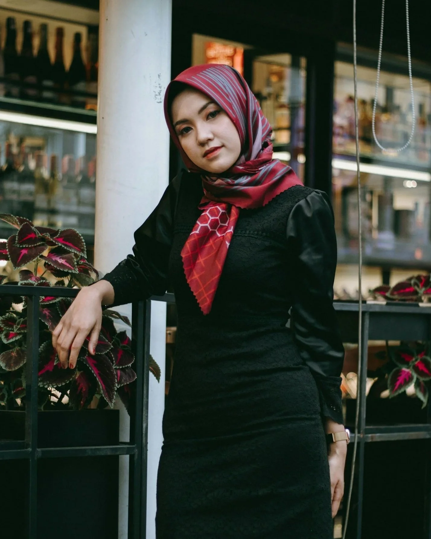 A woman wearing a red hijab and black dress leaning on a metal railing inside a store or mall with plants and shelving in the background.