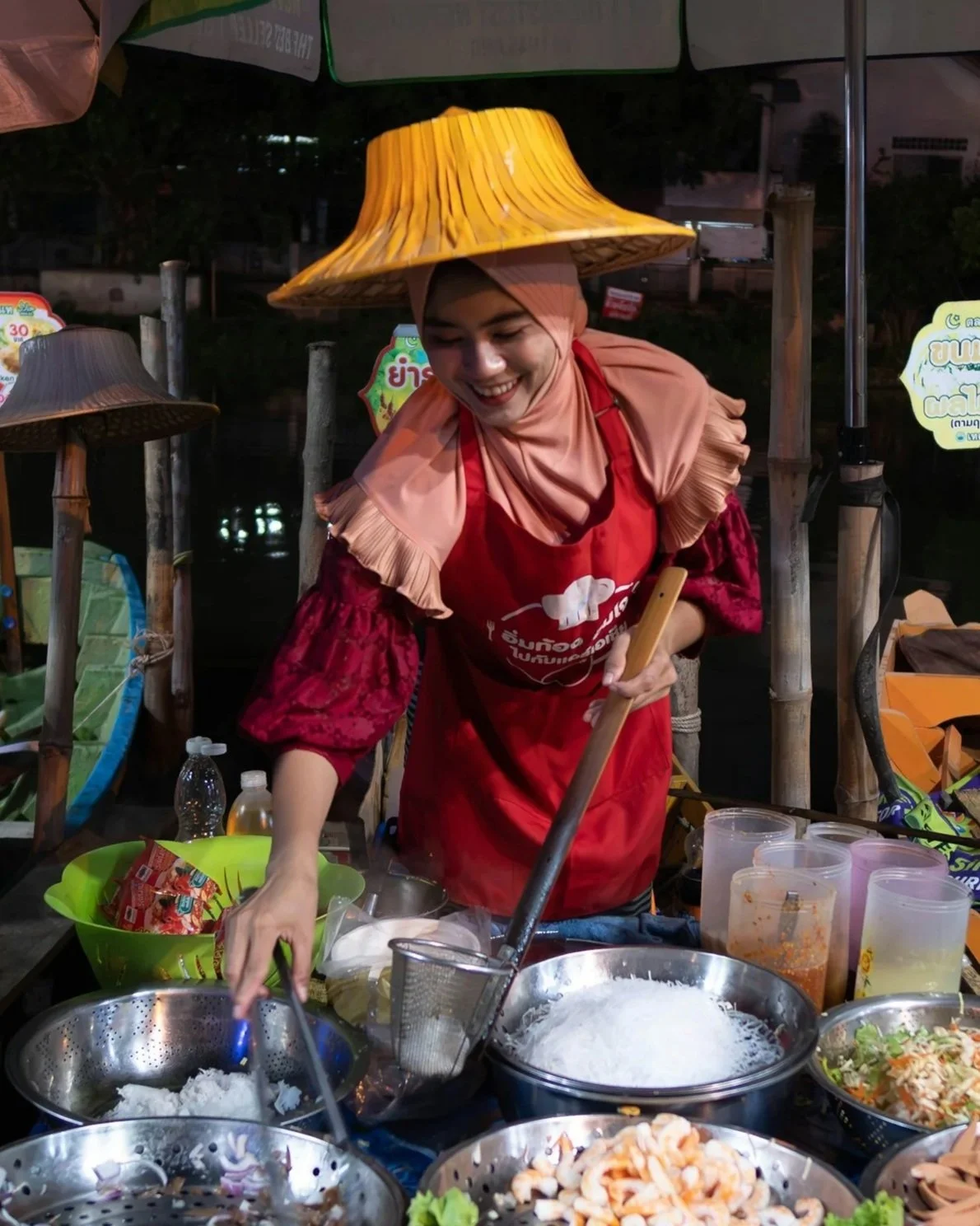 A woman wearing a traditional conical hat and a red apron cooking at an outdoor food stall at night, with various cooking ingredients and utensils on the table.