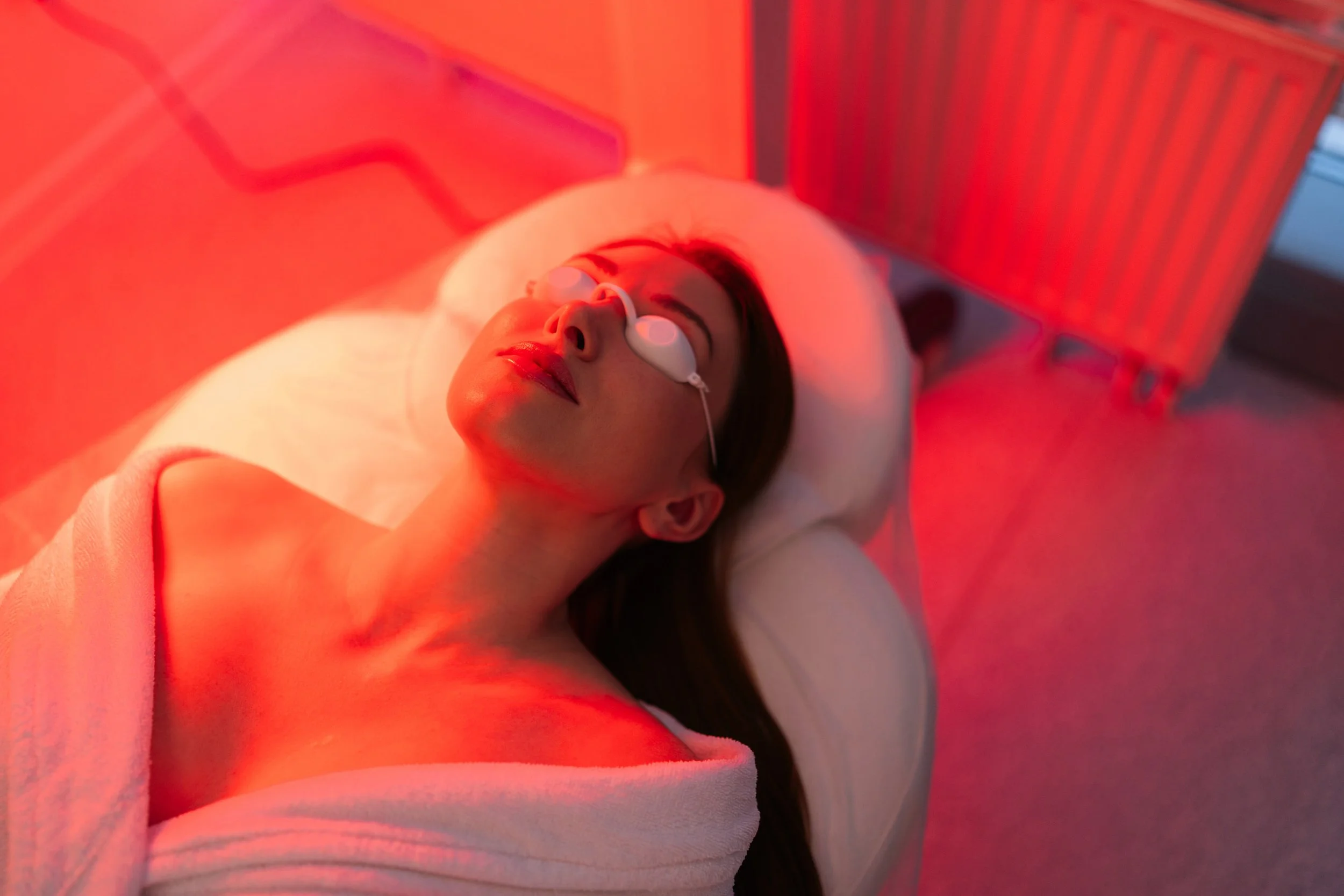 A woman lying on a treatment bed for red light therapy in Nova Scotia with her eyes closed, receiving a facial treatment with a medical device over her eyes, illuminated by red light.