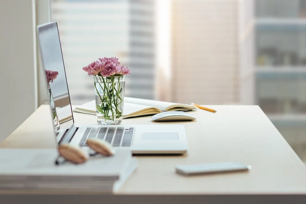 Desk with open laptop, glass vase with pink flowers, notebook, computer mouse, pen, and smartphone in a bright room.