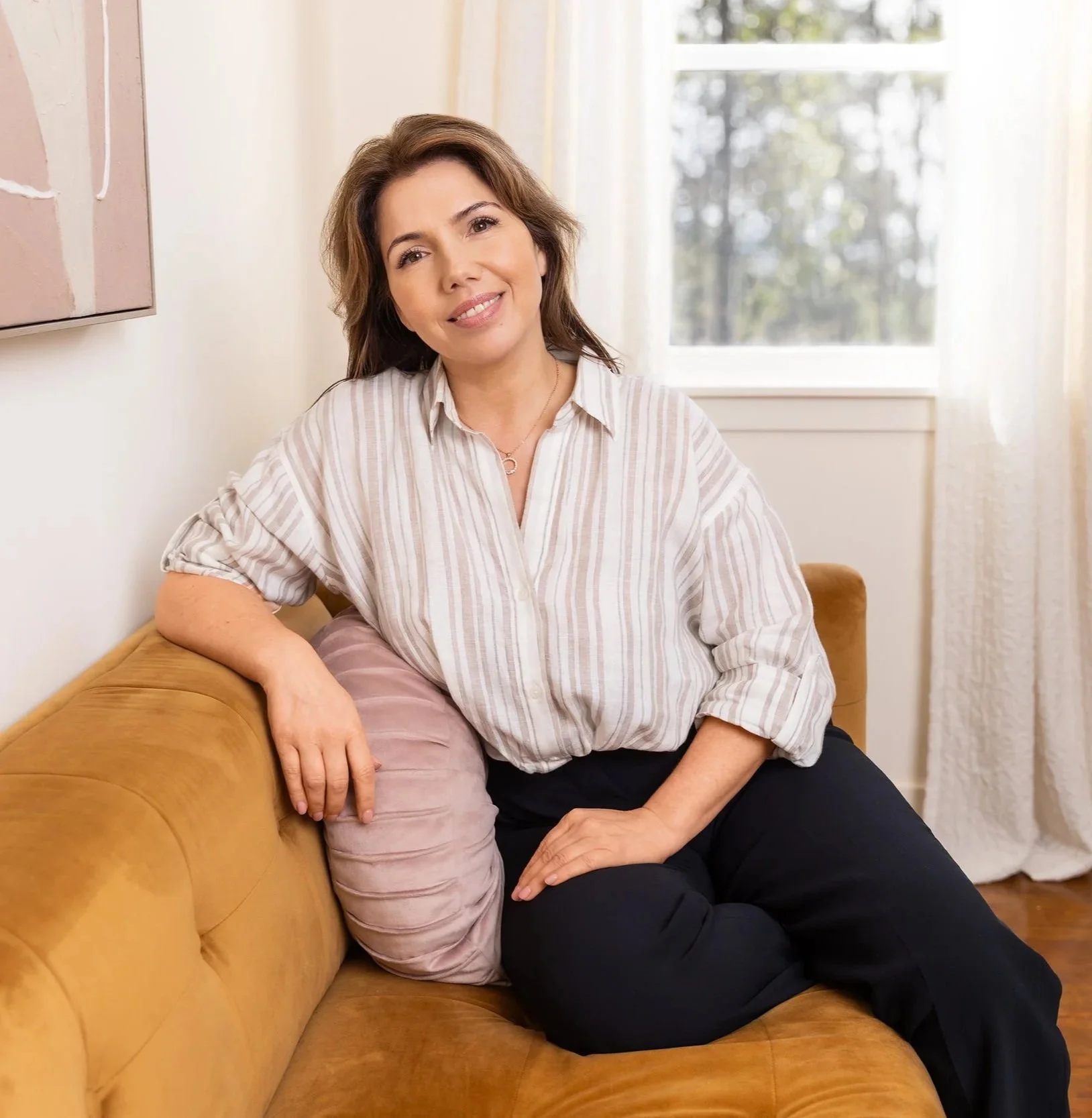 Headshot of Shannon Chappelle Nova Scota Spa owner sitting on a tan sofa in a bright room with white curtains and a window in the background.