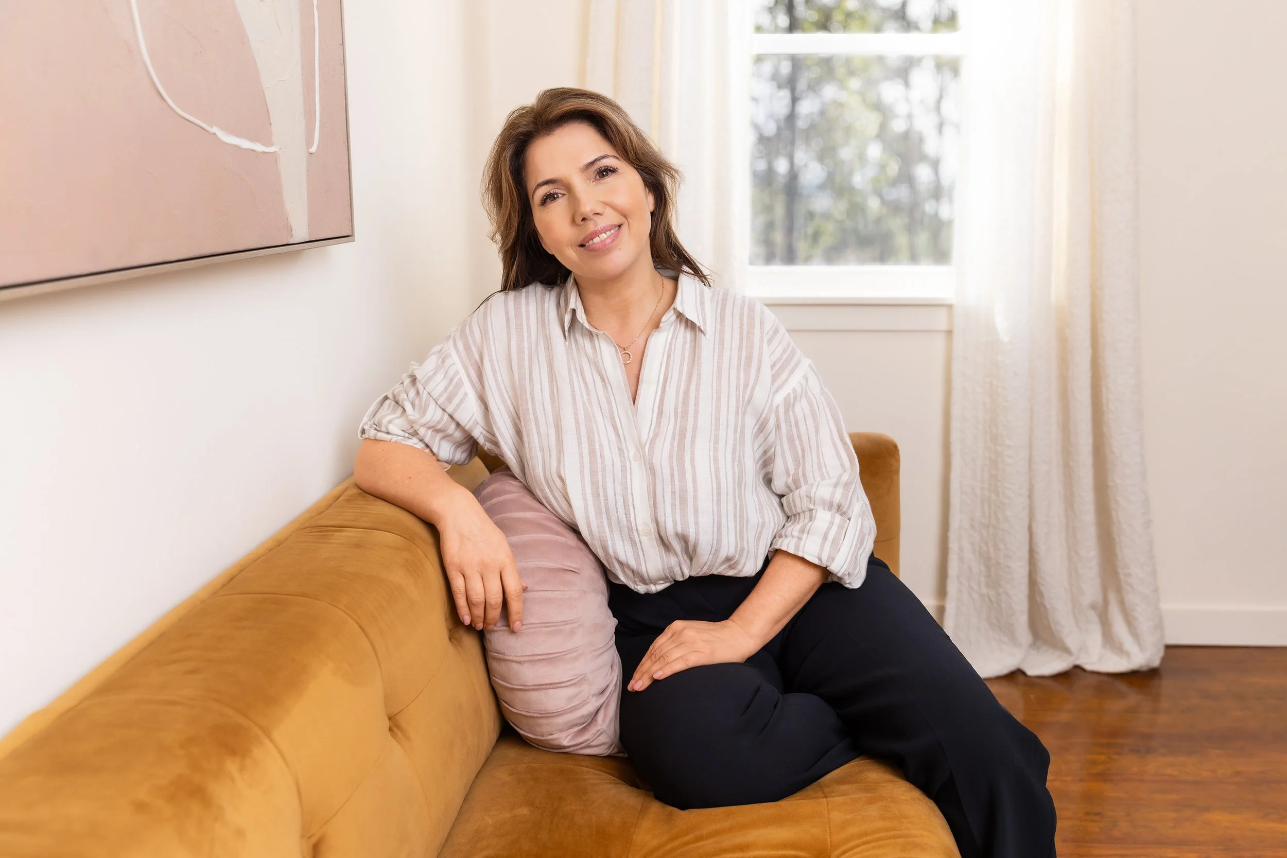 Headshot of Shannon Chappelle Nova Scota Spa owner sitting on a tan sofa in a bright room with white curtains and a window in the background.