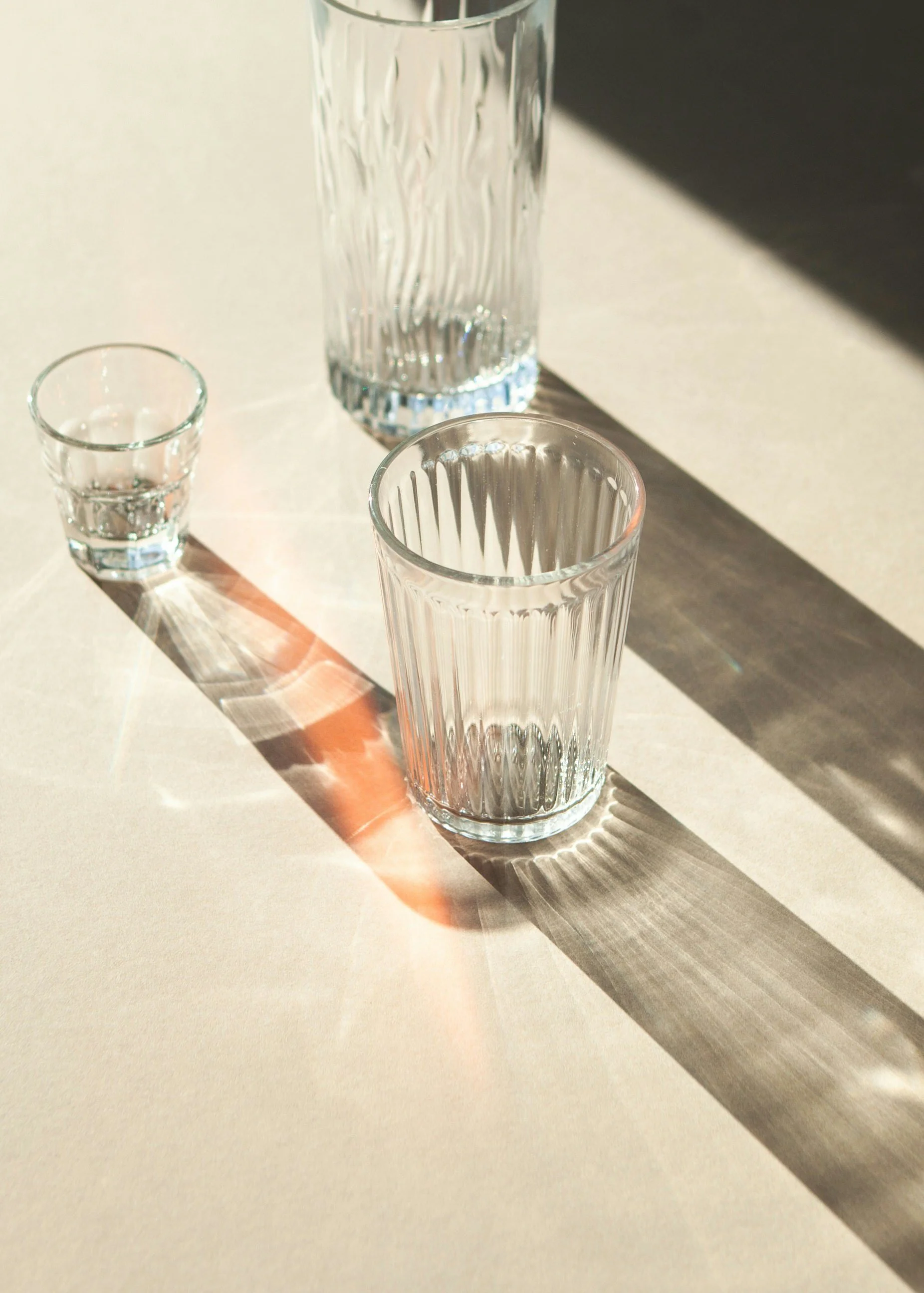 Three glass containers of varying sizes with water, casting colorful shadows and reflections on a light-colored surface.