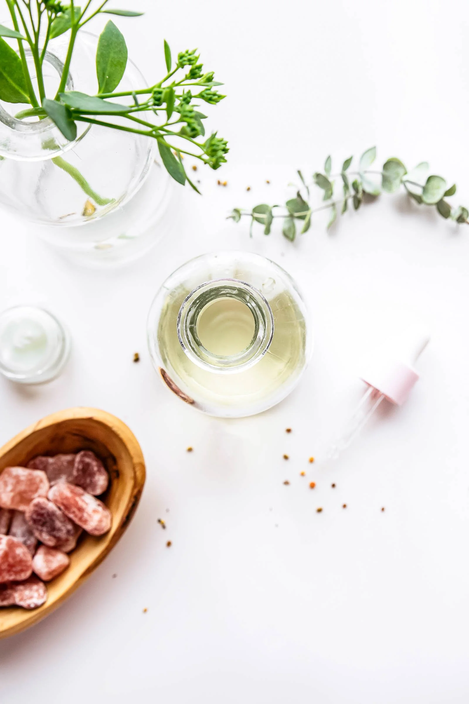A flat lay of a white surface with a glass bottle of essential oil, a small pink dropper, a wooden bowl of pink Himalayan salt rocks, and a few sprigs of green herbs.