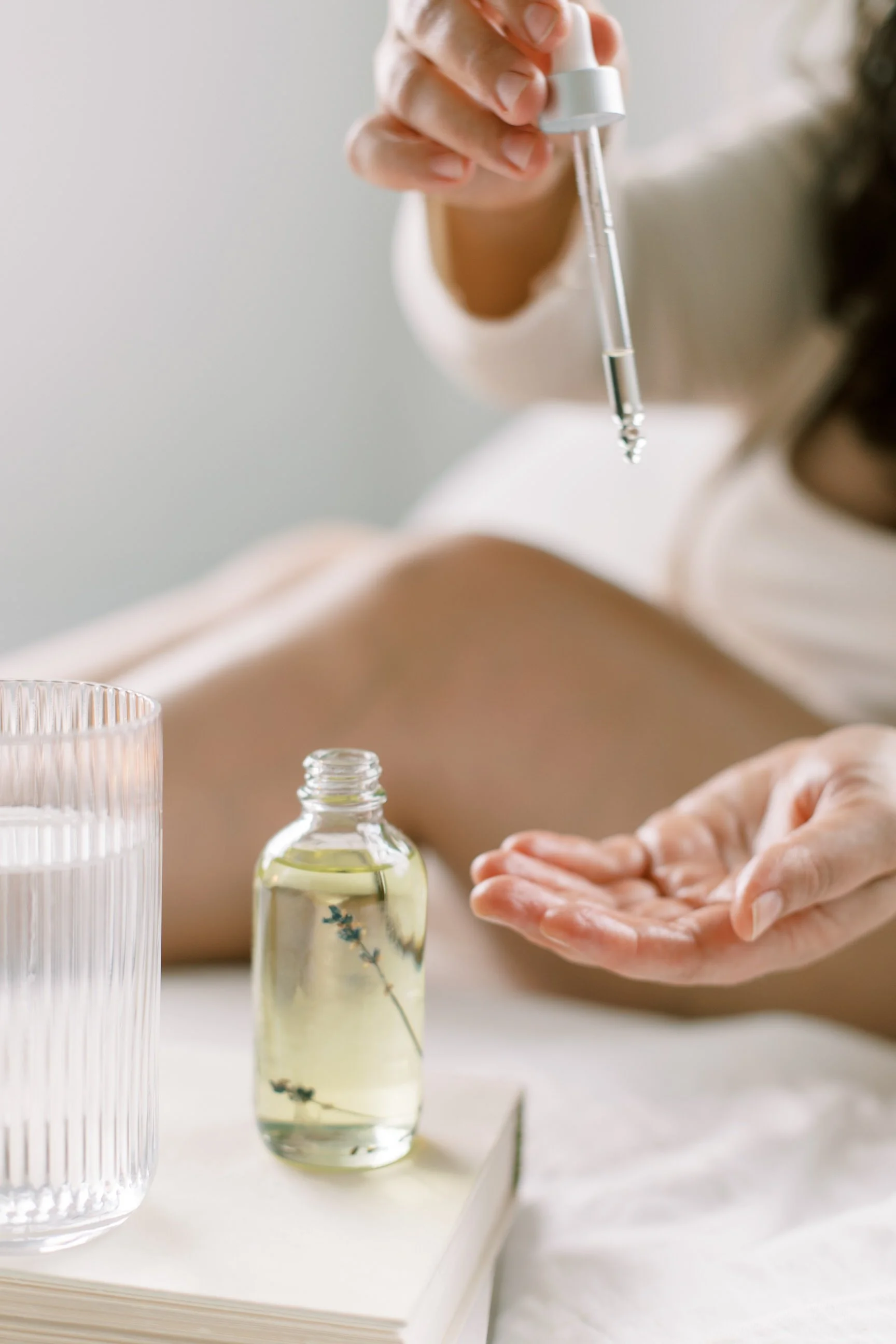 Person administering facial serum with dropper, with a bottle of oil and glass of water nearby.