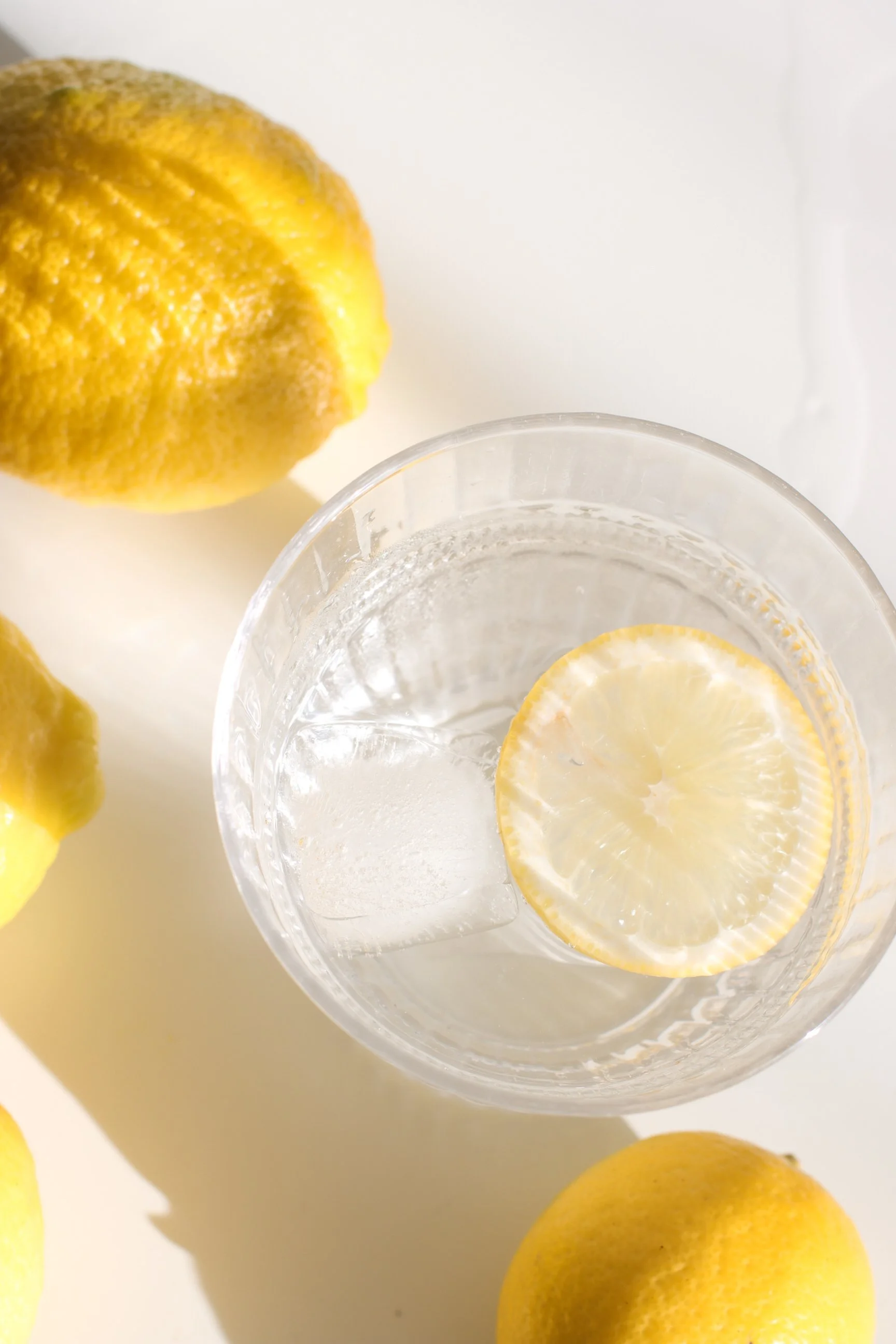A glass of water with a lemon slice inside, surrounded by whole lemons on a white surface.
