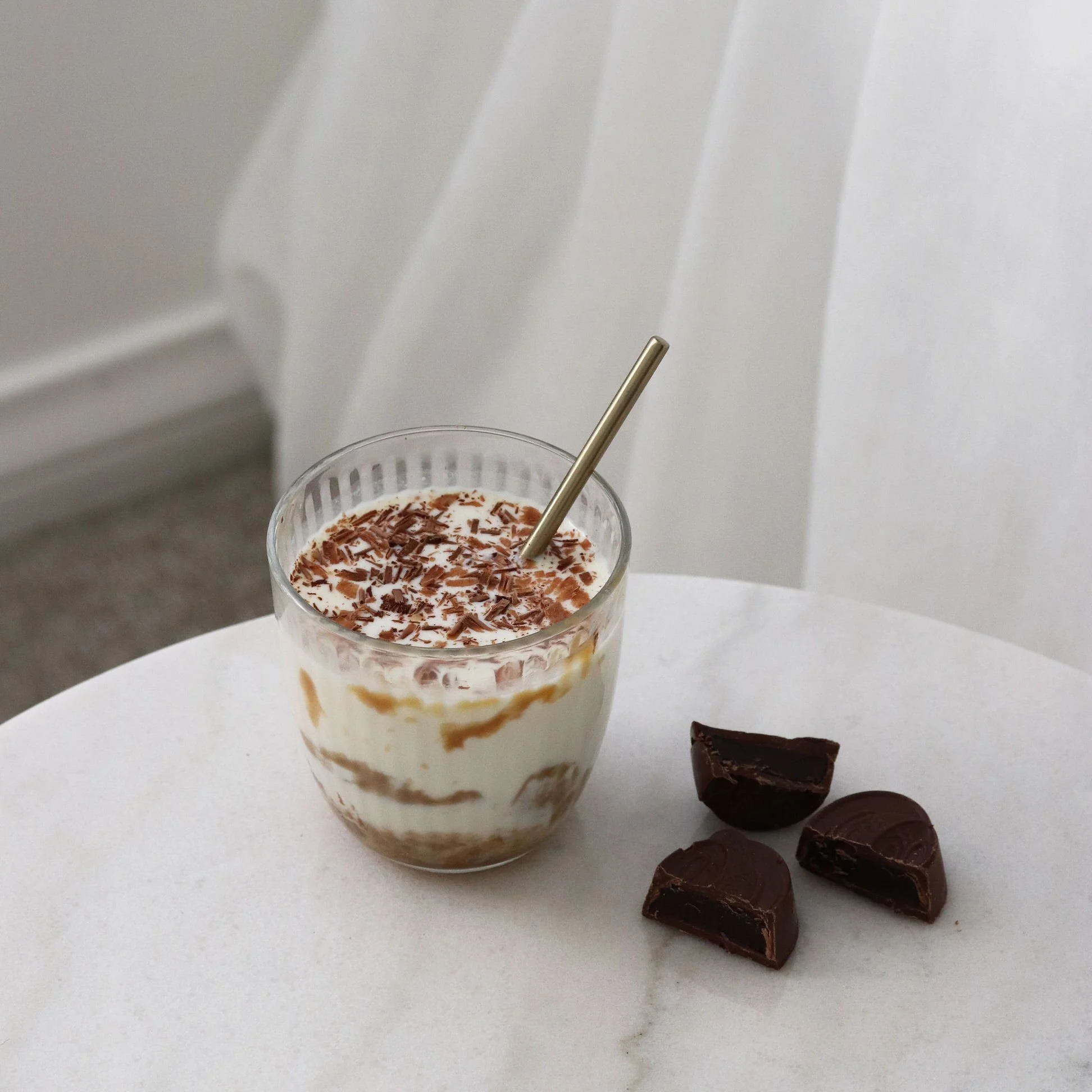 A glass of layered dessert with cream, chocolate shavings, and caramel on top, with three pieces of dark chocolate on a white table.