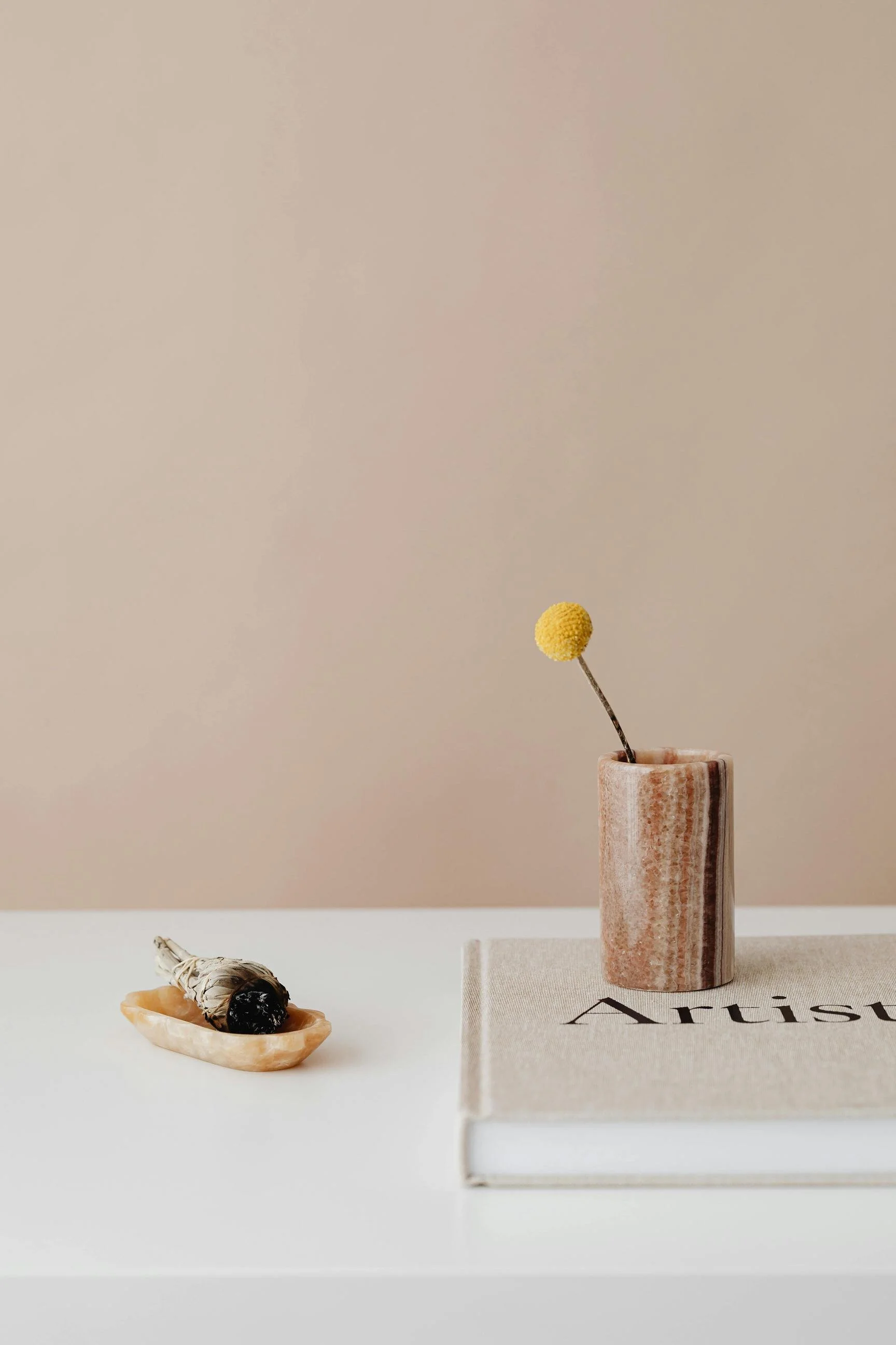 Minimalist styled interior scene features a white surface with a small beige bowl holding a white sage smudge stick, a hardcover art book, and a pinkish marble vase with a single yellow billy button flower. The background has a soft beige wall.