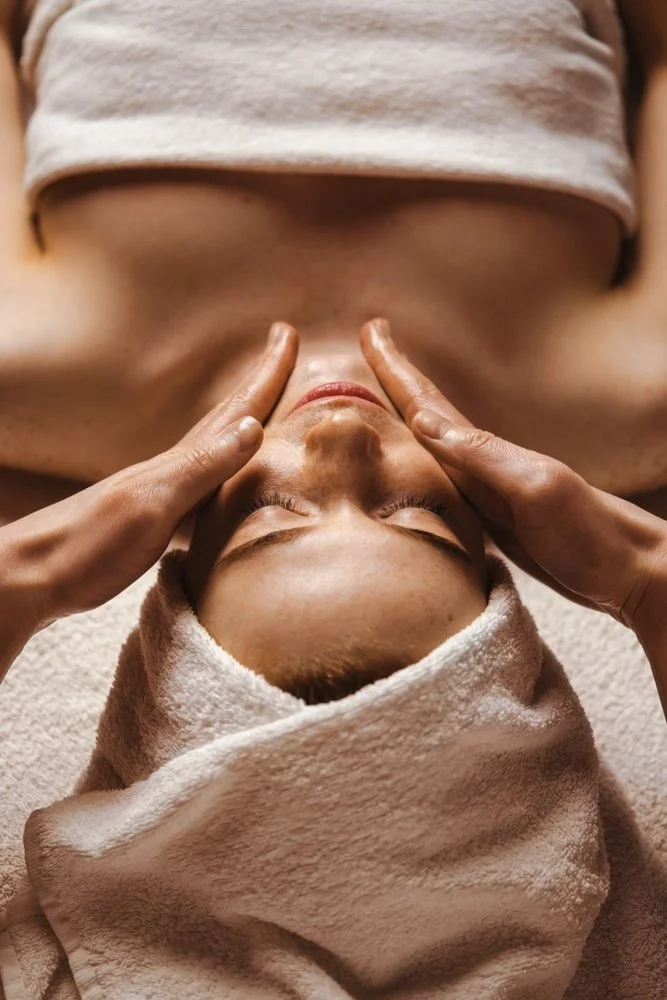 A person receiving a facial massage at a spa, lying on a towel with a towel wrapped around their head.