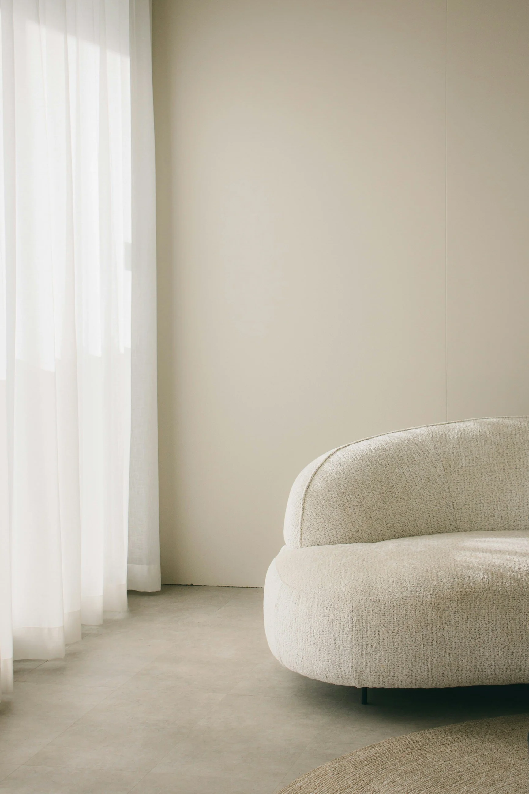 A minimalist living room corner with white curtains, a beige curved sofa, and matching beige carpet.