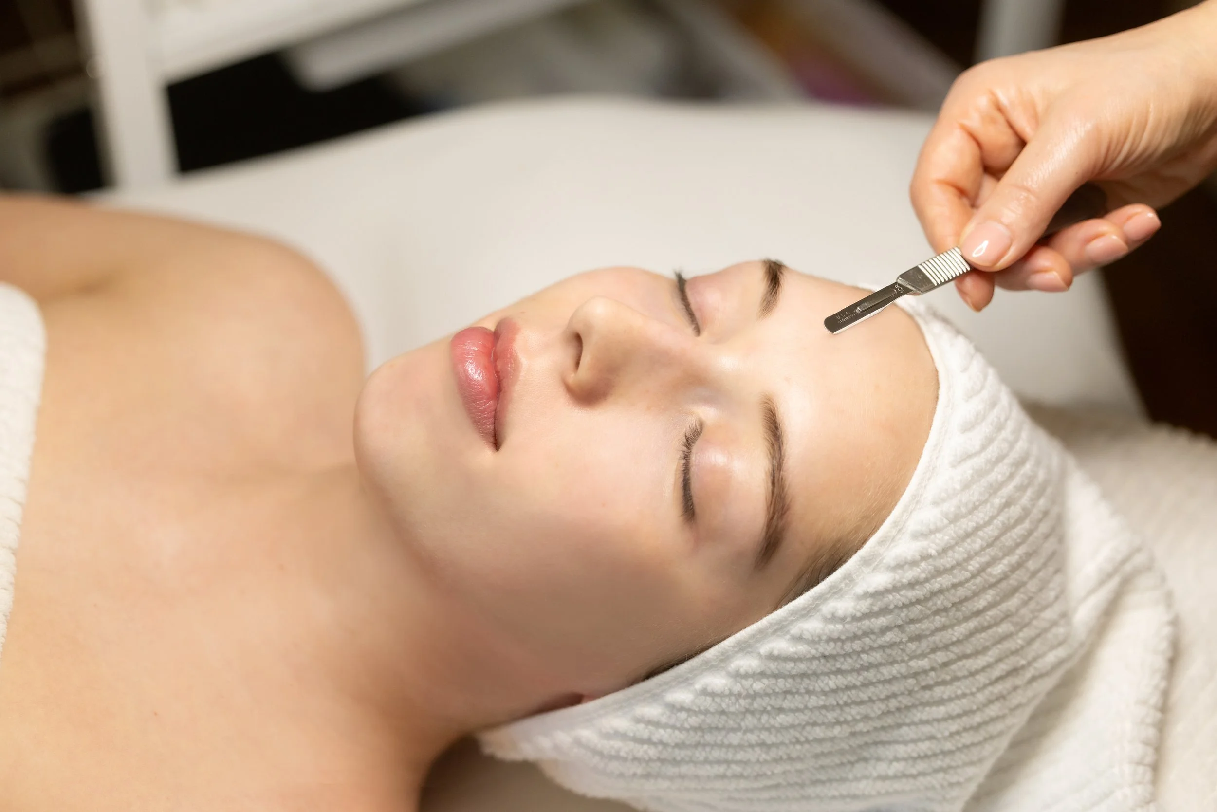 A woman receiving a facial treatment with a small metal blade on her forehead in a spa setting.