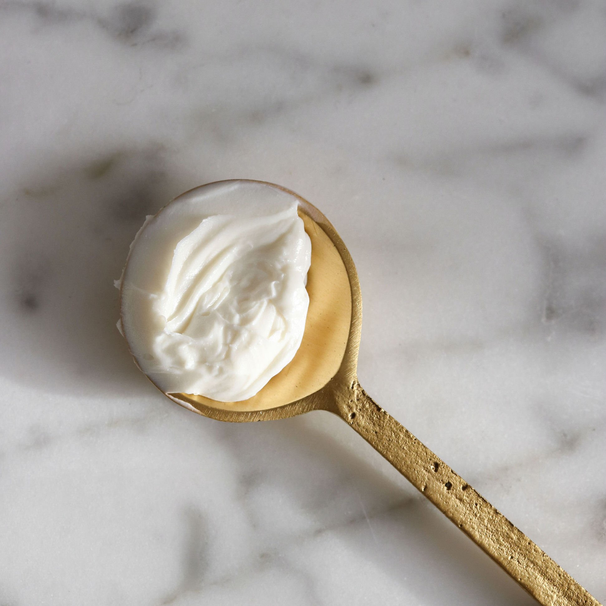 A gold spoon filled with a dollop of white cream or yogurt, placed on a white marble surface.