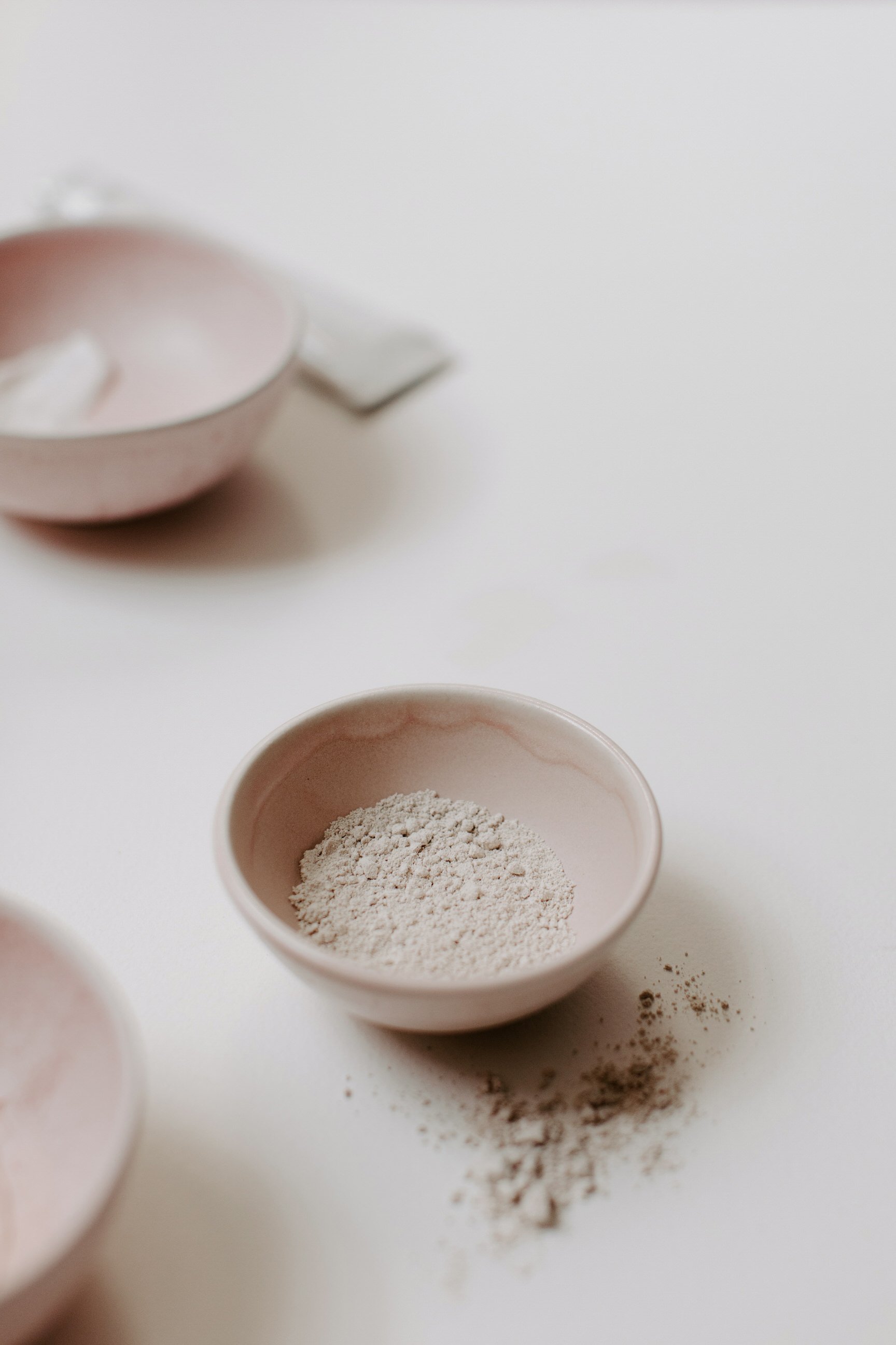 A small pink bowl filled with white powder, with some powder spilled on the white surface next to it; a second pink bowl with more white powder is blurred in the background.
