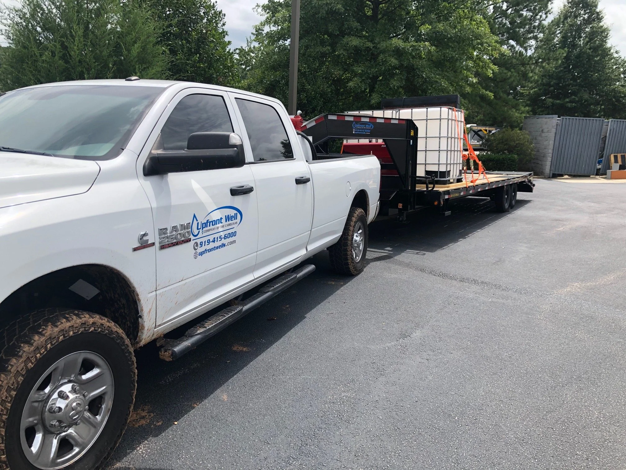 White pickup truck with company logo parked in an outdoor lot with a flatbed trailer carrying large containers and equipment.