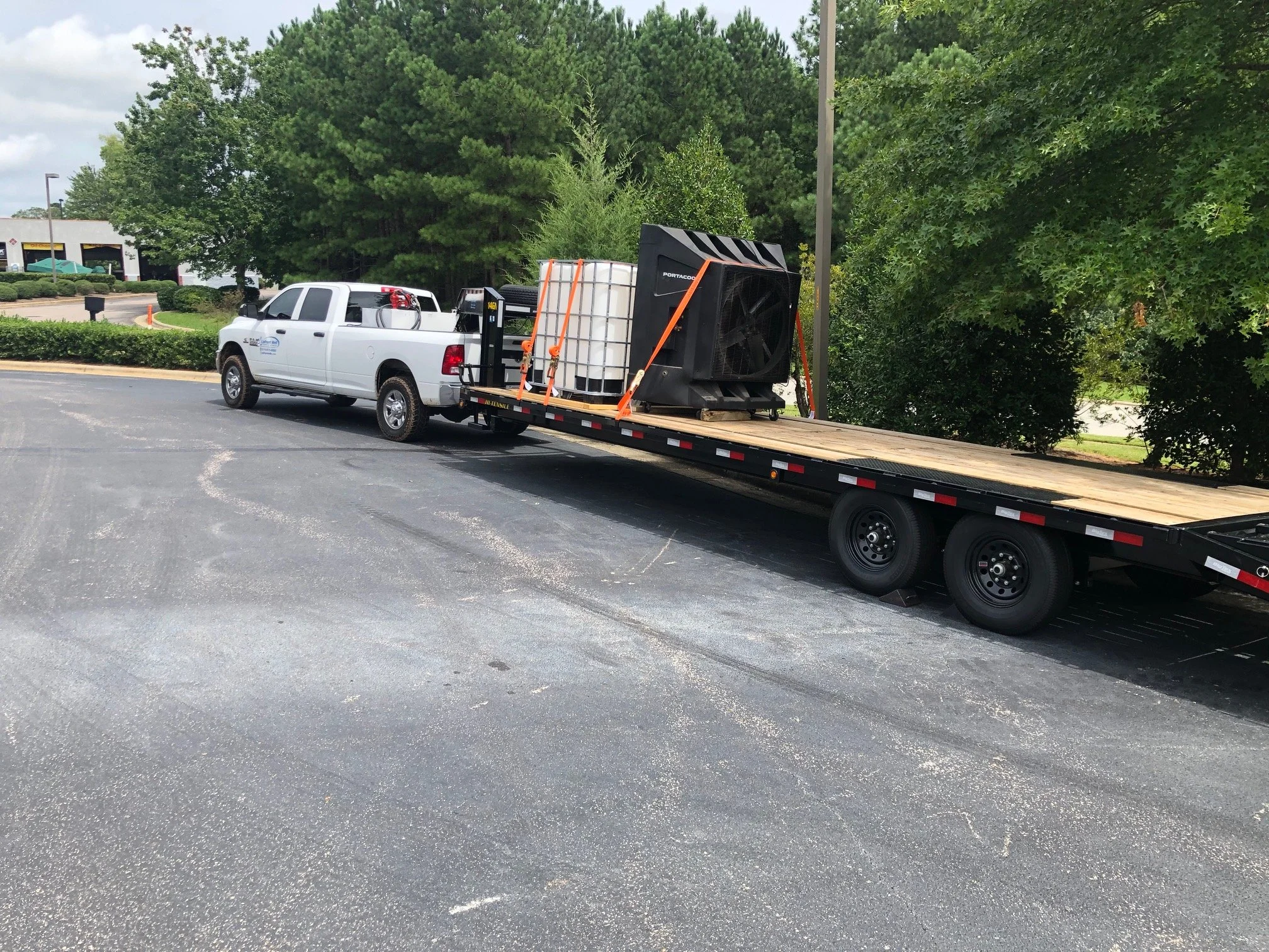 A white pickup truck hauling a flatbed trailer with a large air conditioning unit and a water tank, parked on a paved lot with green trees in the background.
