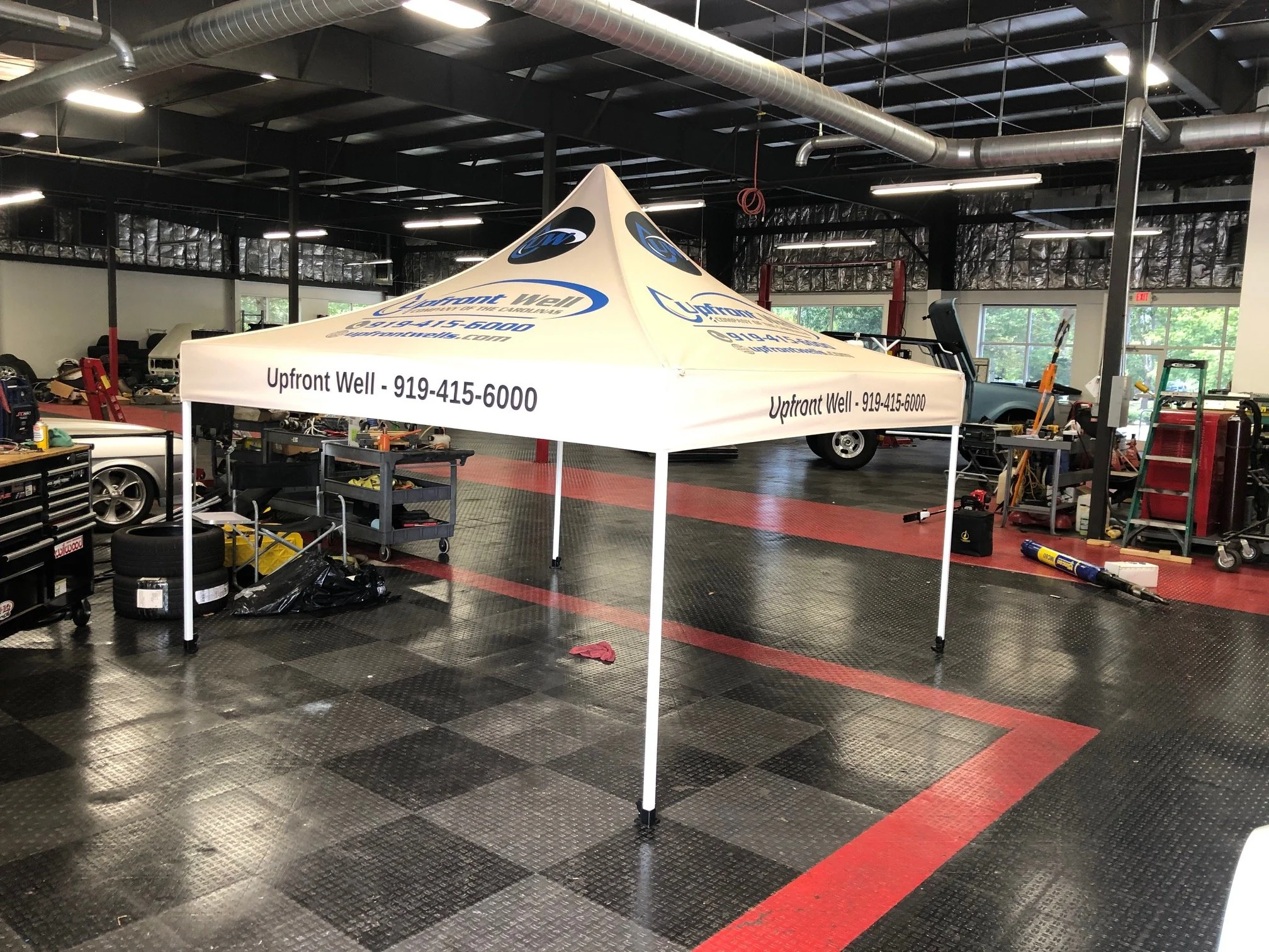 A white canopy tent with the text 'Upfront Well - 919-415-6000' printed on it, set up inside a garage or workshop with tools, cars, and equipment around.