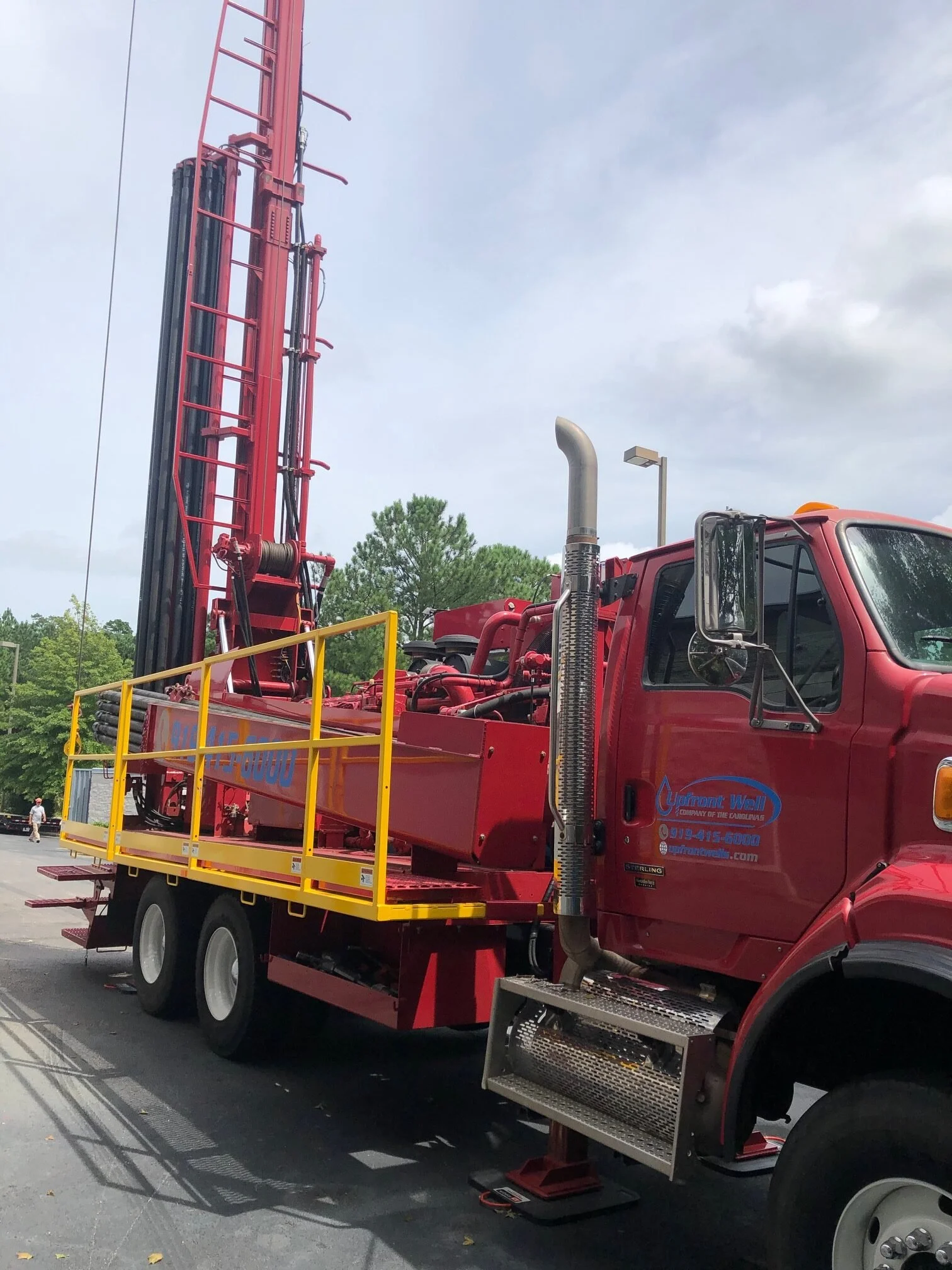 A red drilling truck with a tall, vertical drilling rig on a parking lot, with trees and partly cloudy sky in the background.