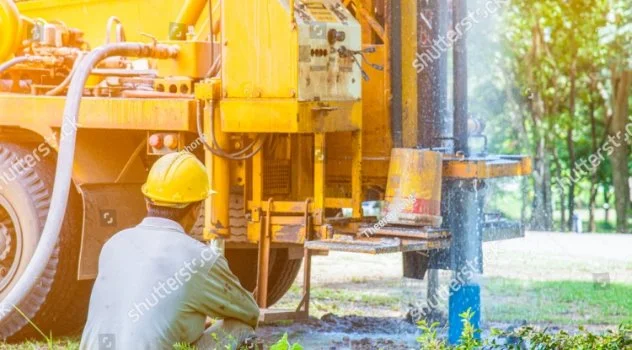 Worker in a yellow helmet operating drilling equipment outdoors in a park or green area.