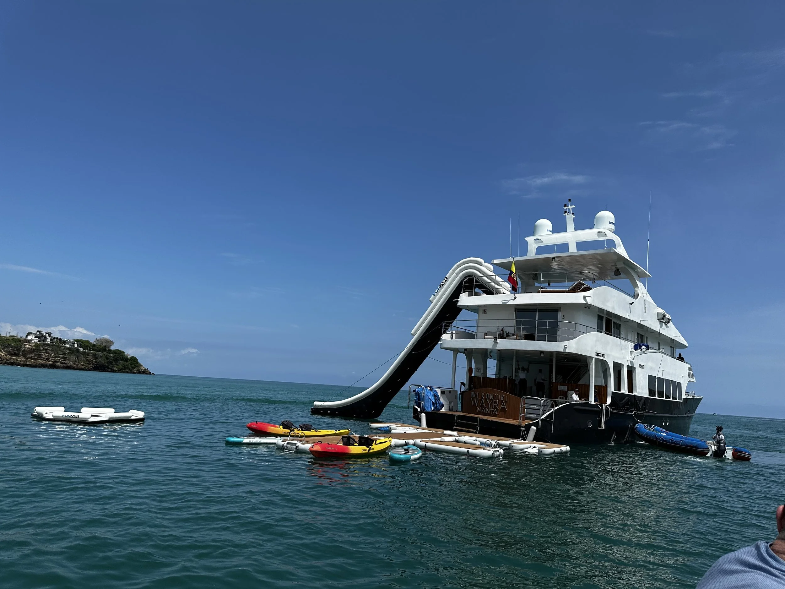 Large white yacht docked near a rocky shoreline, with a small white dinghy nearby, and several colorful paddleboards and kayaks floating in the water, under a clear blue sky.